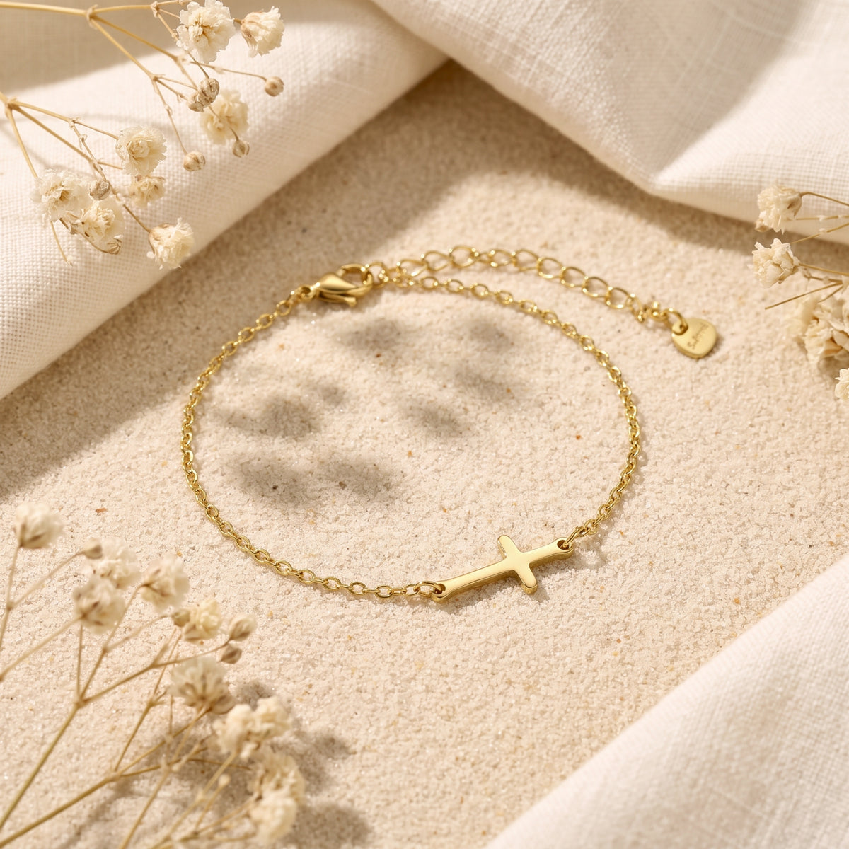 Gold-tone chain bracelet with an integrated horizontal cross, displayed flat on sand beside white fabric and dried flowers.