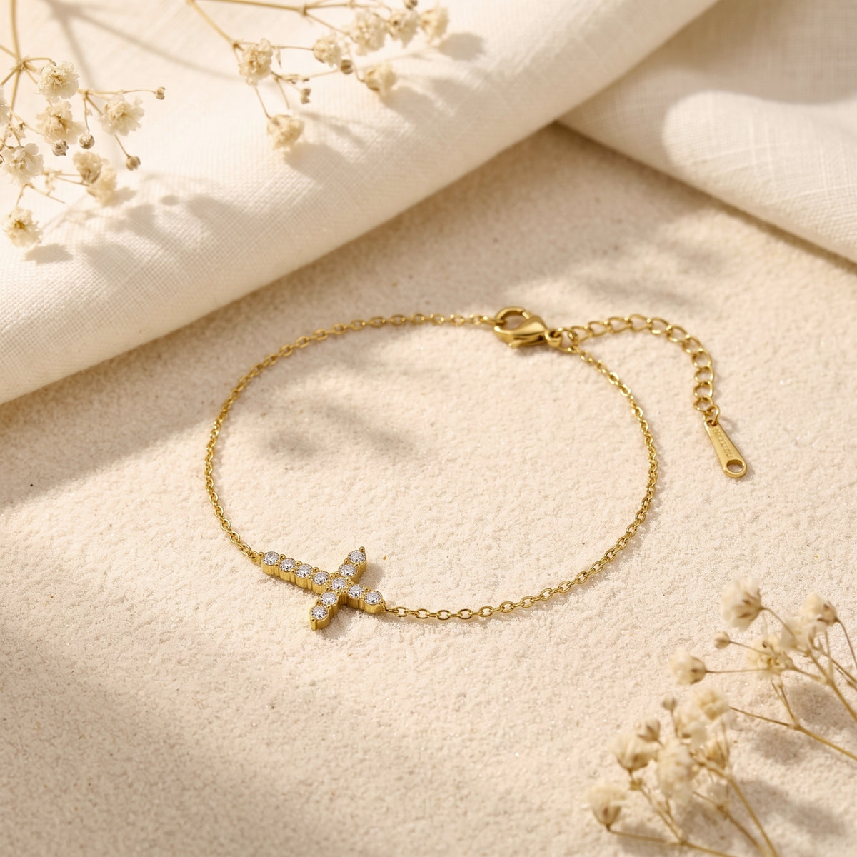 Gold-tone chain bracelet with a cubic zirconia-set sideways cross resting on textured sand and white linen.