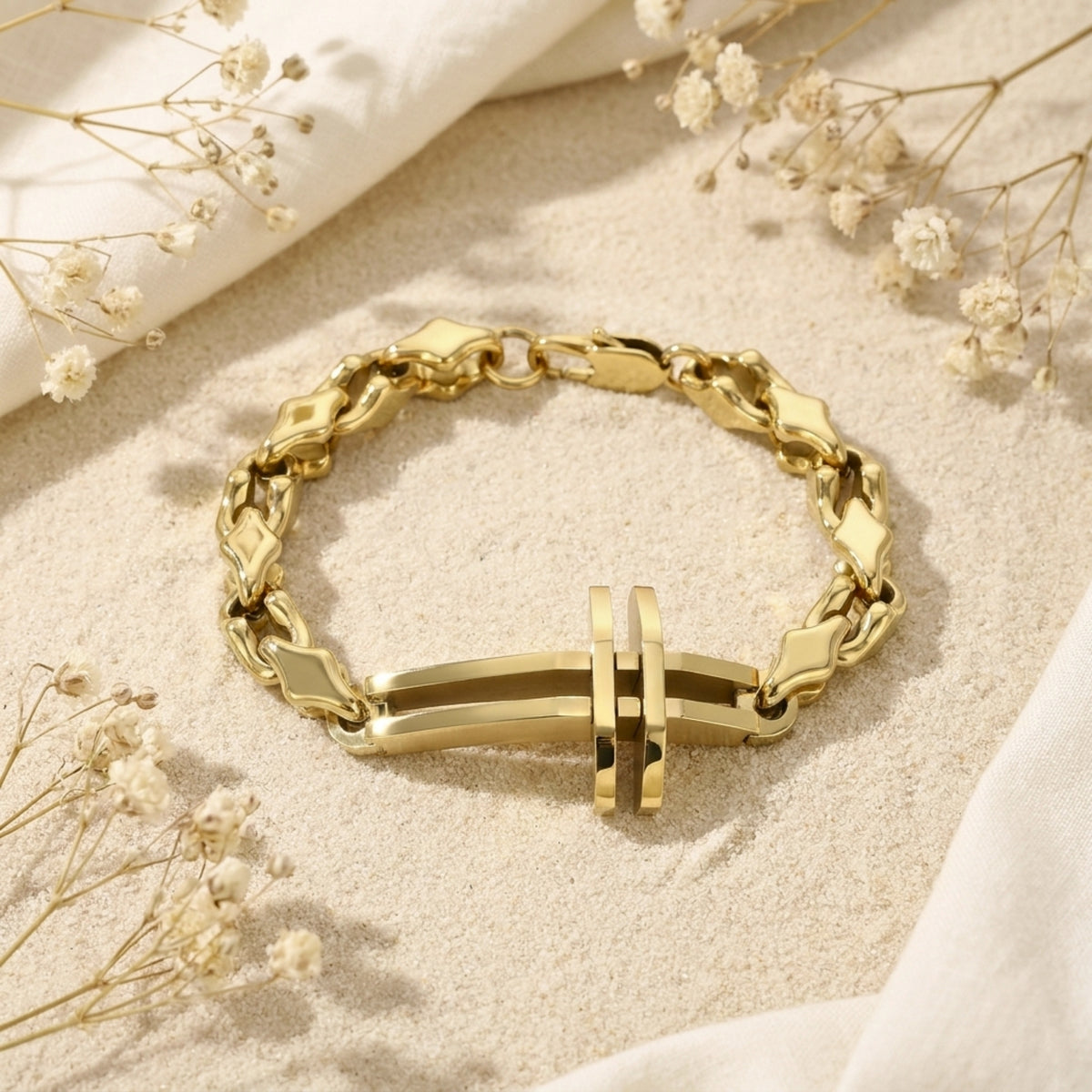 Gold-tone sculpted cross link bracelet laid on sand with dried white flowers.