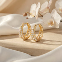 Gold round-cut moissanite J-hoop earrings on a stone surface with white orchids and silk.