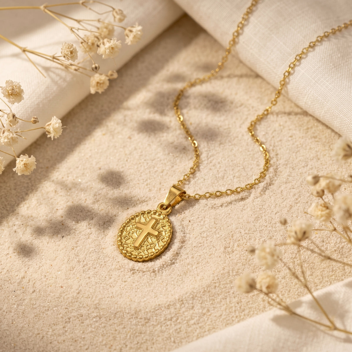 Gold-tone cross medallion necklace lying on sand with dried flowers and white fabric