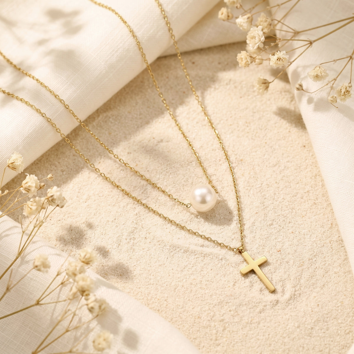 Gold-tone layered necklace with a white pearl station and cross pendant, arranged on sand with dried flowers.
