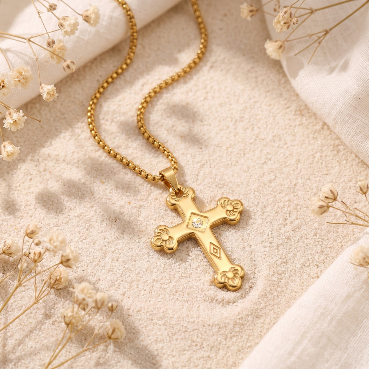 Gold-tone floriated cross pendant with a clear central accent lying on sand among white dried flowers.