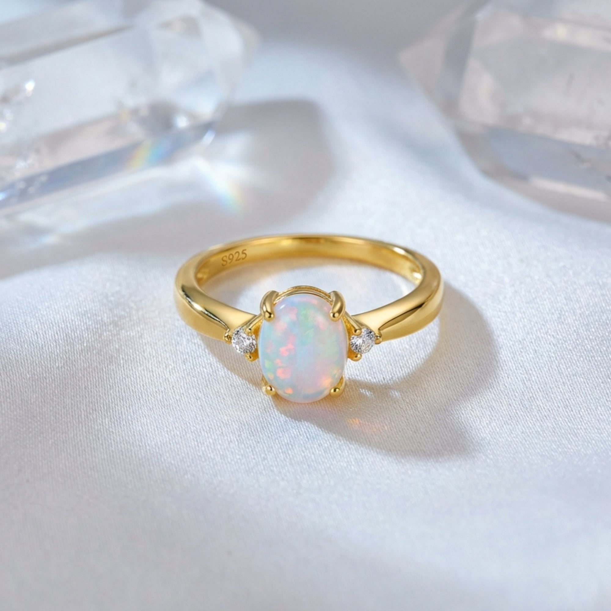 Gold-tone S925 silver ring with a natural oval opal and side stones, styled on white silk with crystals.