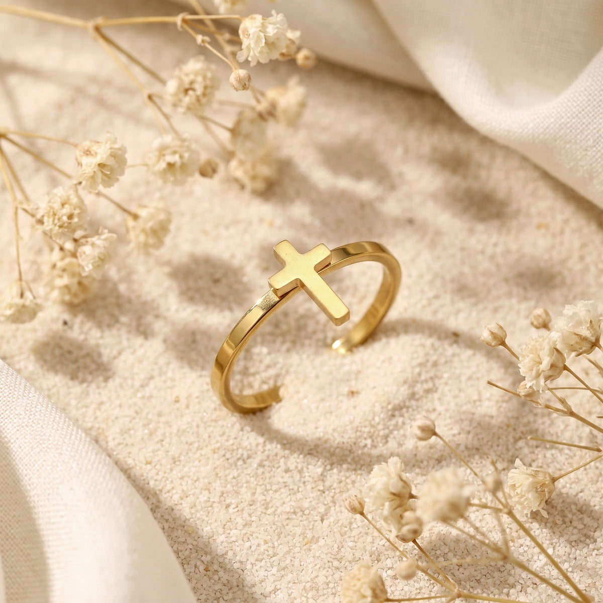 Gold-tone adjustable cross ring displayed on fine sand with dried white floral accents.