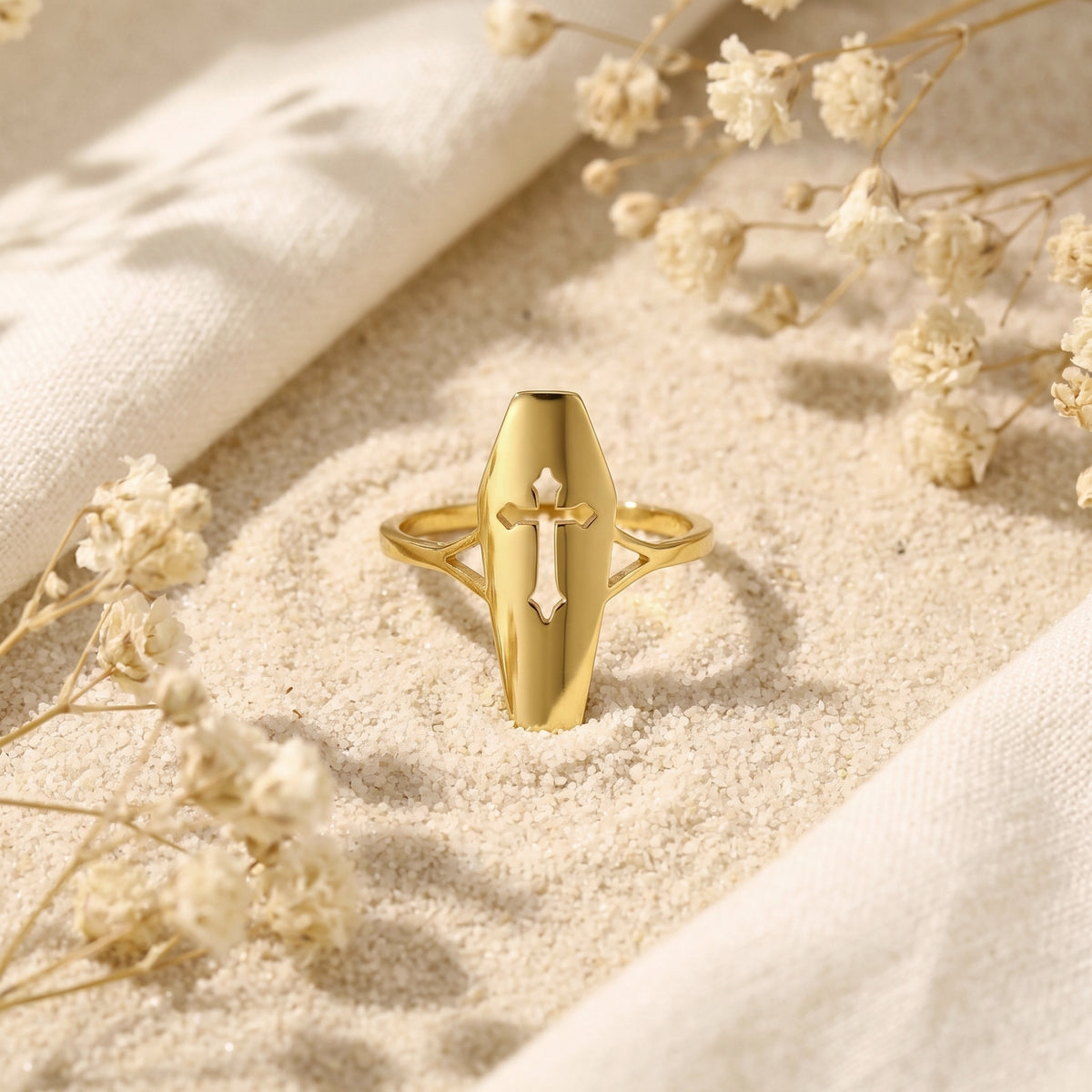 Front view of a gold-tone coffin ring with a cutout cross standing upright in fine sand.