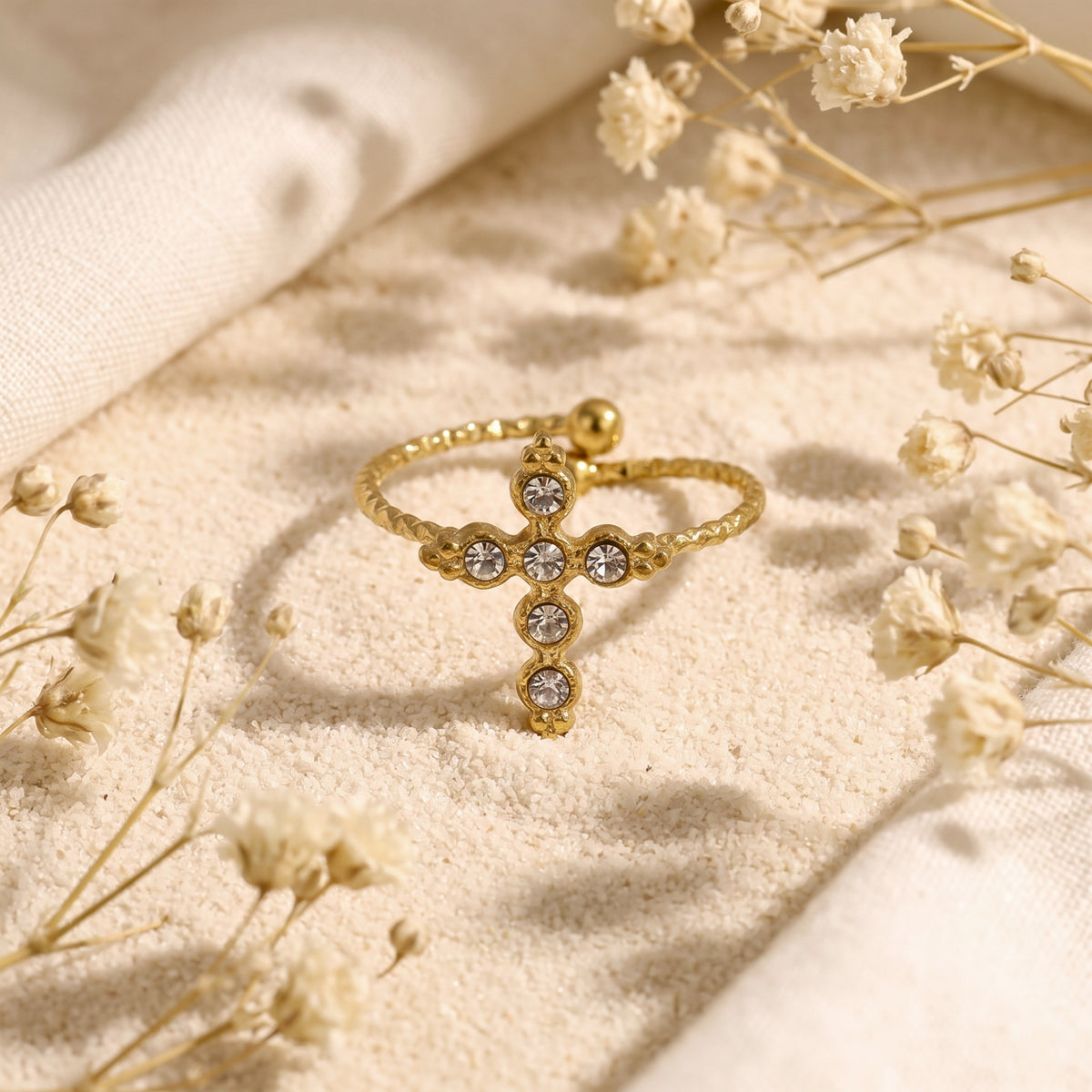 Gold-tone cross ring with clear crystals resting on fine sand among dried flowers.