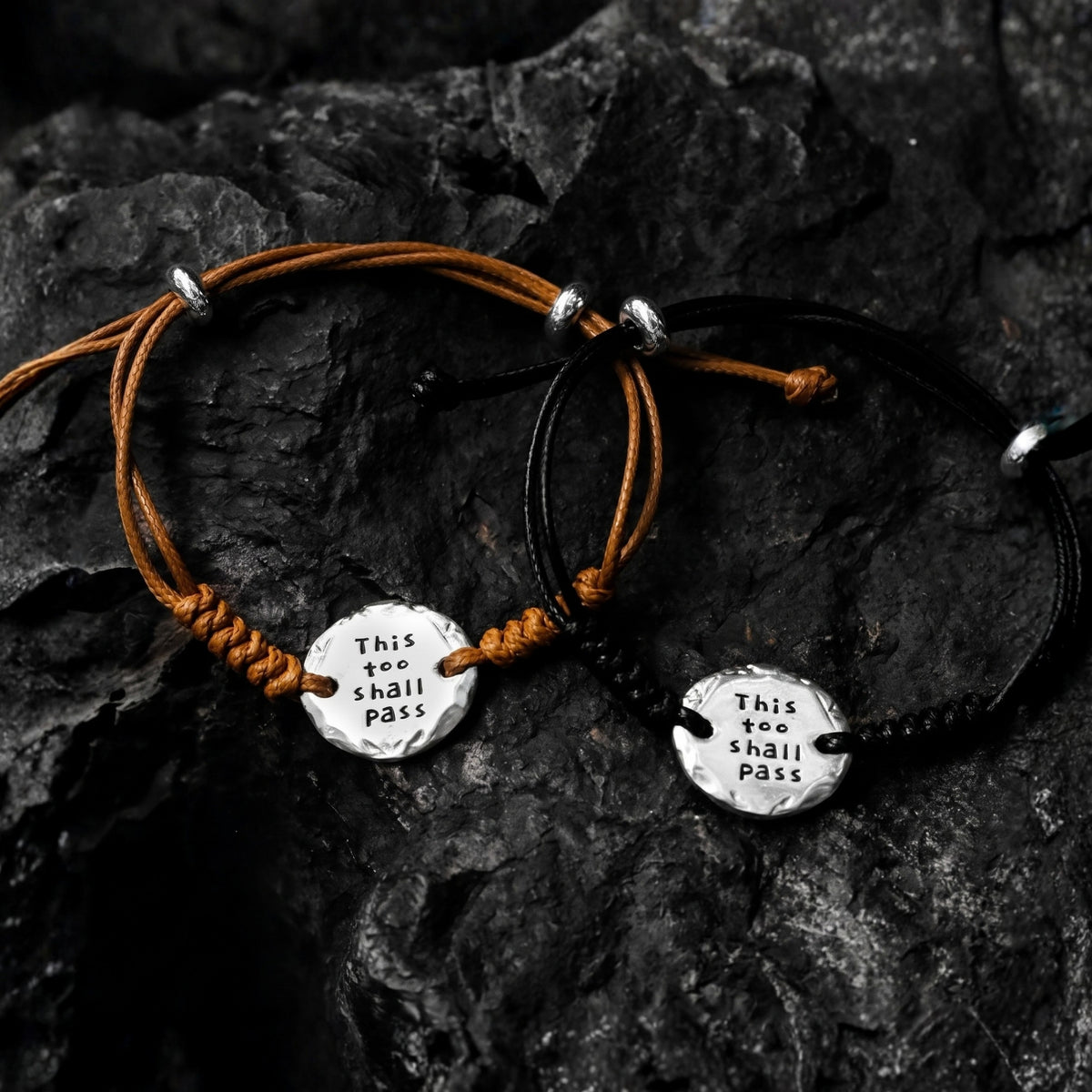 Two brown and black cord bracelets on dark rock, each with a silver medallion stamped “THIS TOO SHALL PASS.”