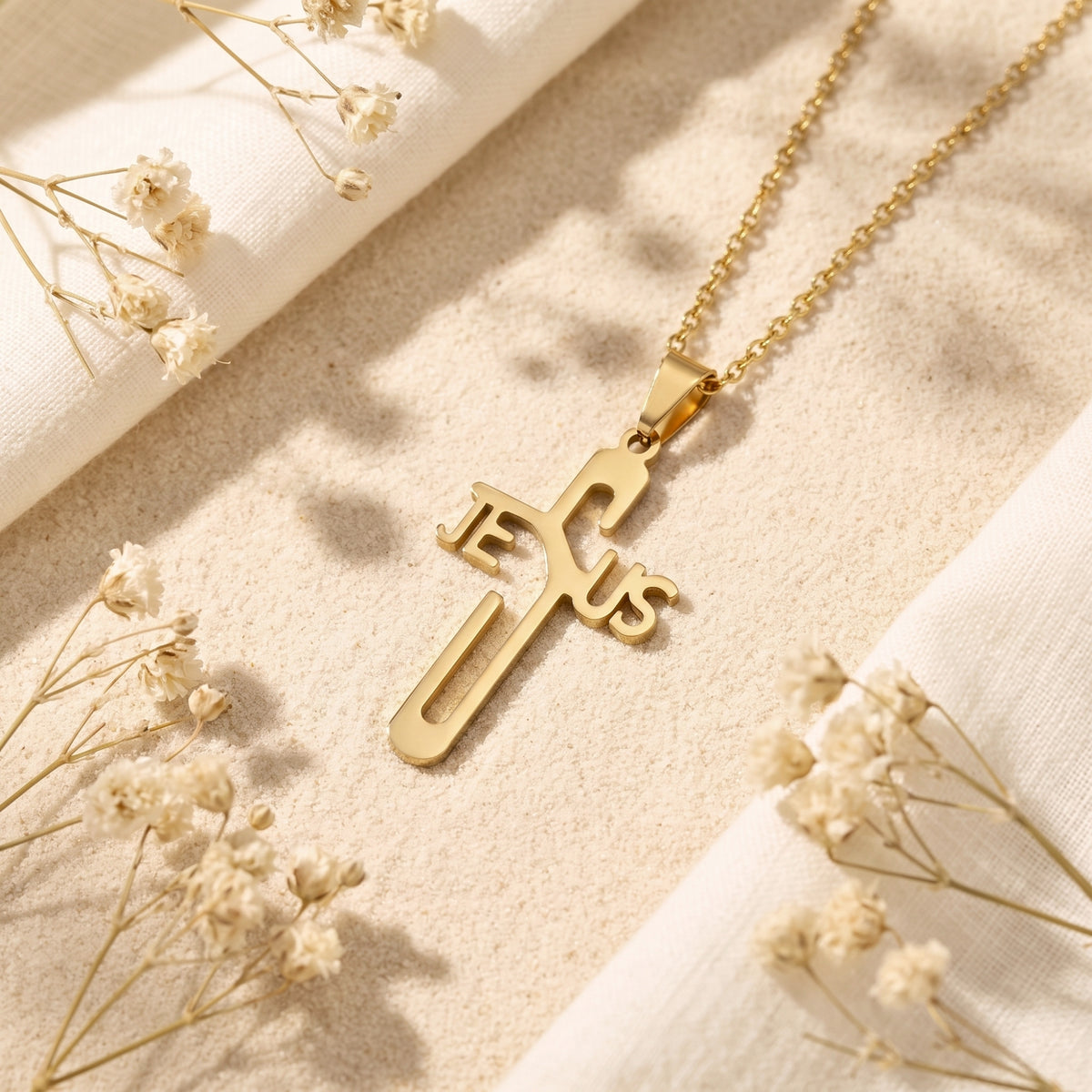 Gold-tone Jesus cross necklace lying on sand beside cream fabric and dried flowers.