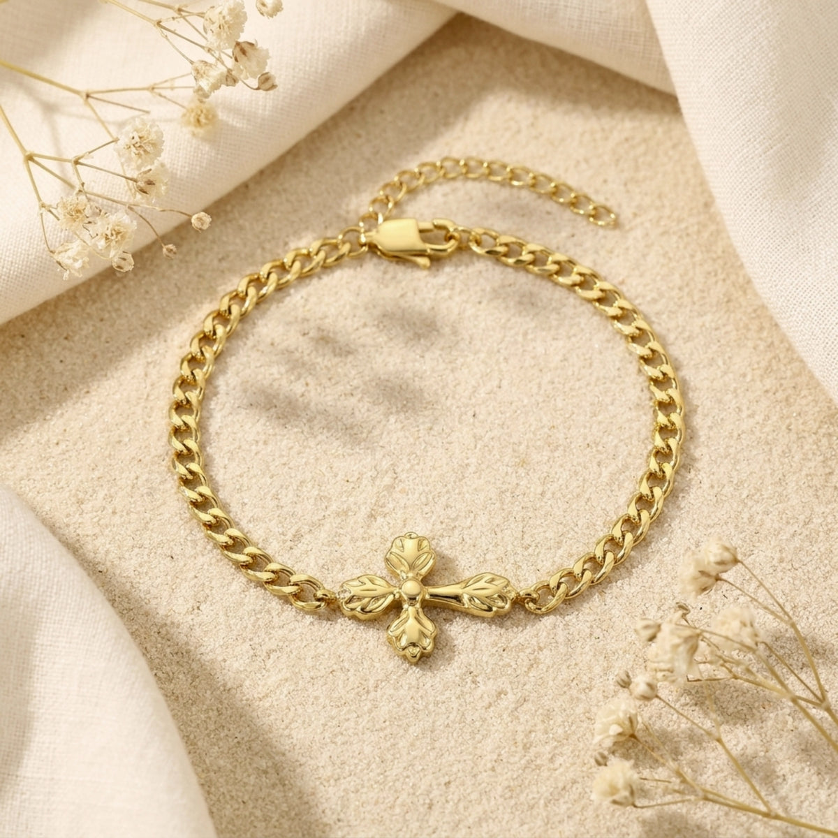 Gold-tone curb chain bracelet with an ornate cross centerpiece laid flat on a sandy surface with dried flowers.