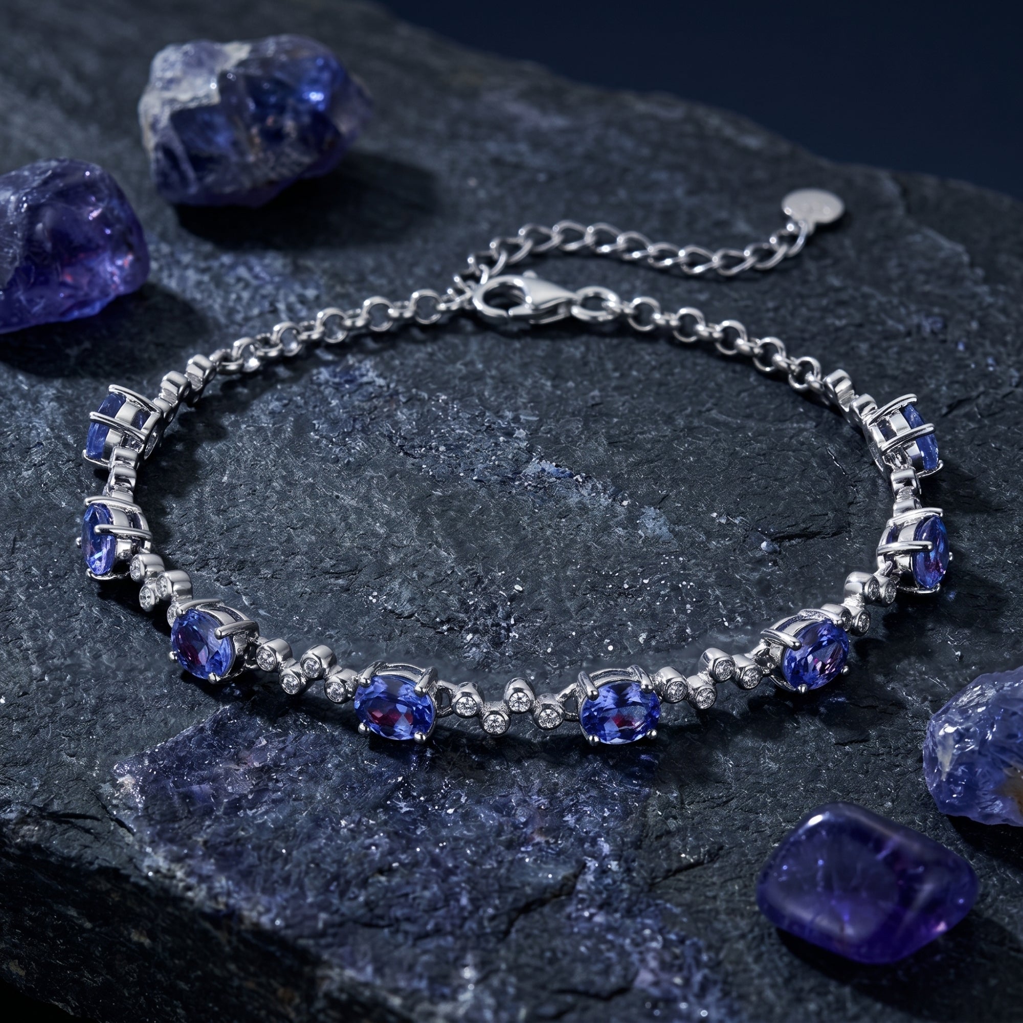 Oval cut natural tanzanite station bracelet in sterling silver on a dark stone background with raw gems.