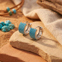Sterling silver huggie earrings with turquoise cabochons displayed on a desert rock with sand and fabric.