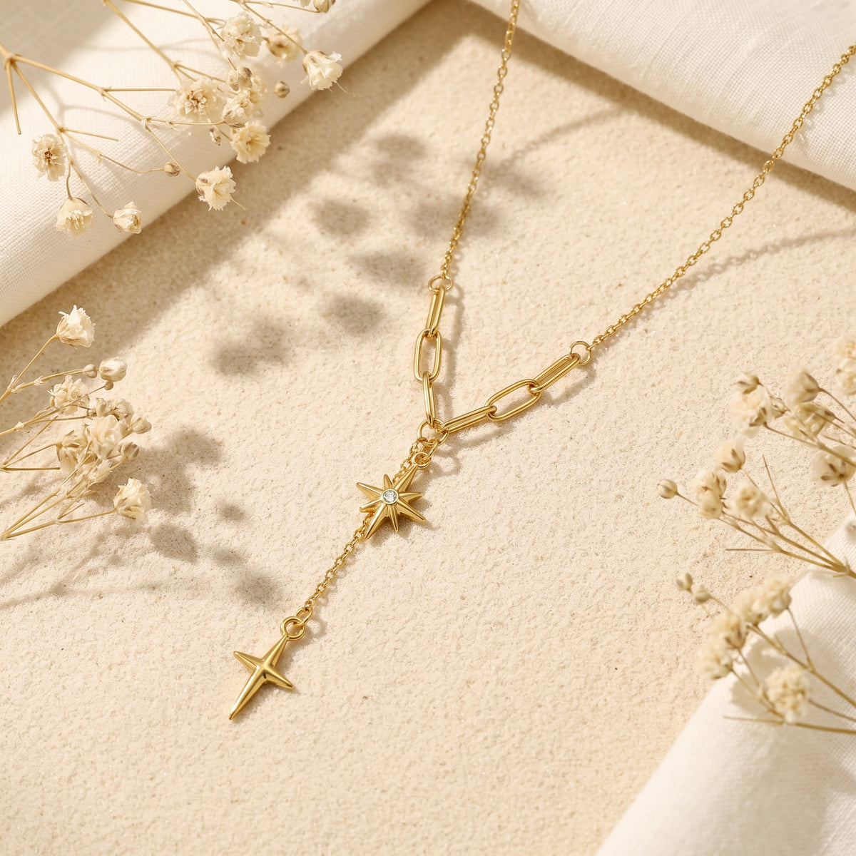 Gold-tone starburst lariat necklace resting diagonally on sand, framed by off-white fabric and white dried flowers.