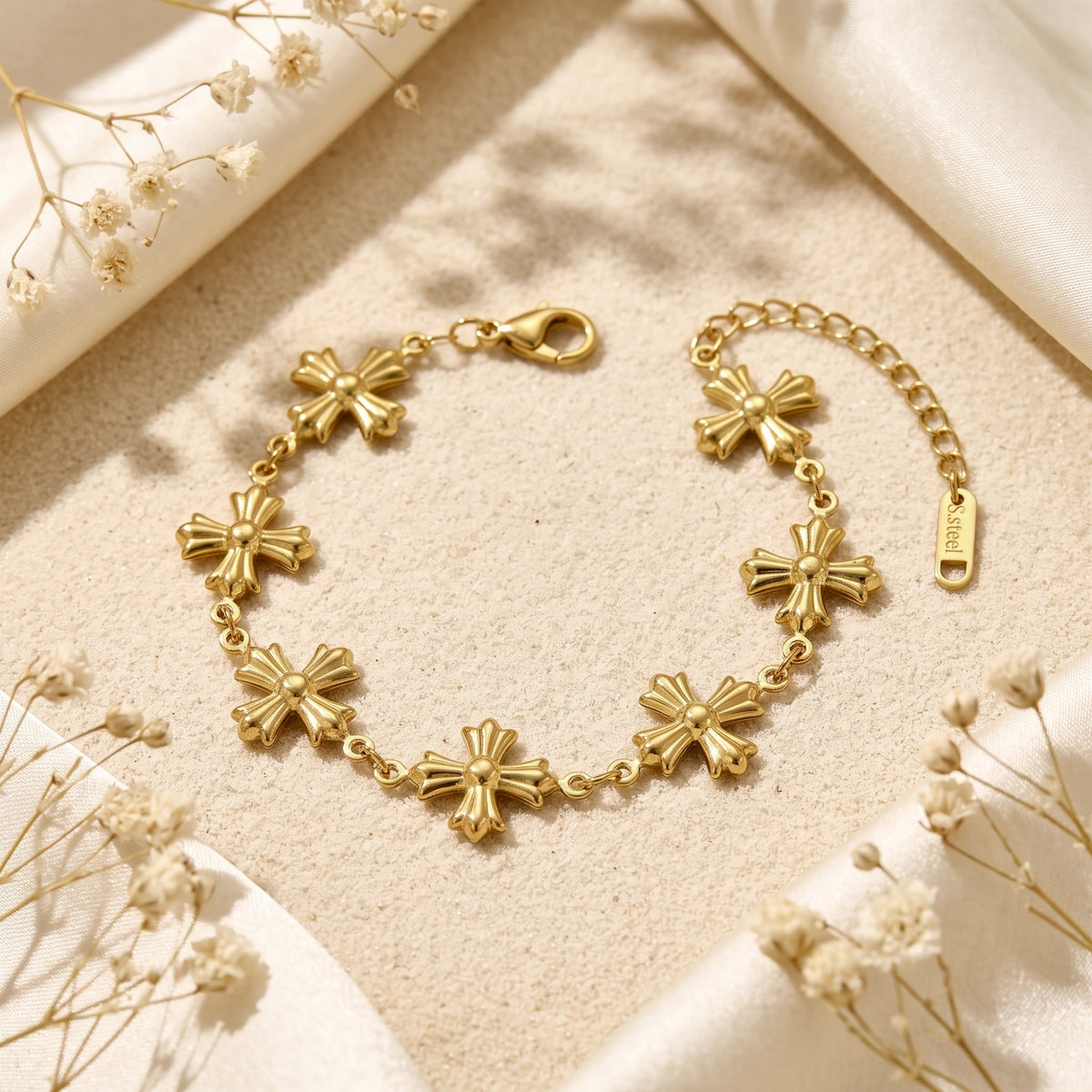 Gold sculpted cross station bracelet displayed flat on sand, framed by cream fabric and dried flowers.
