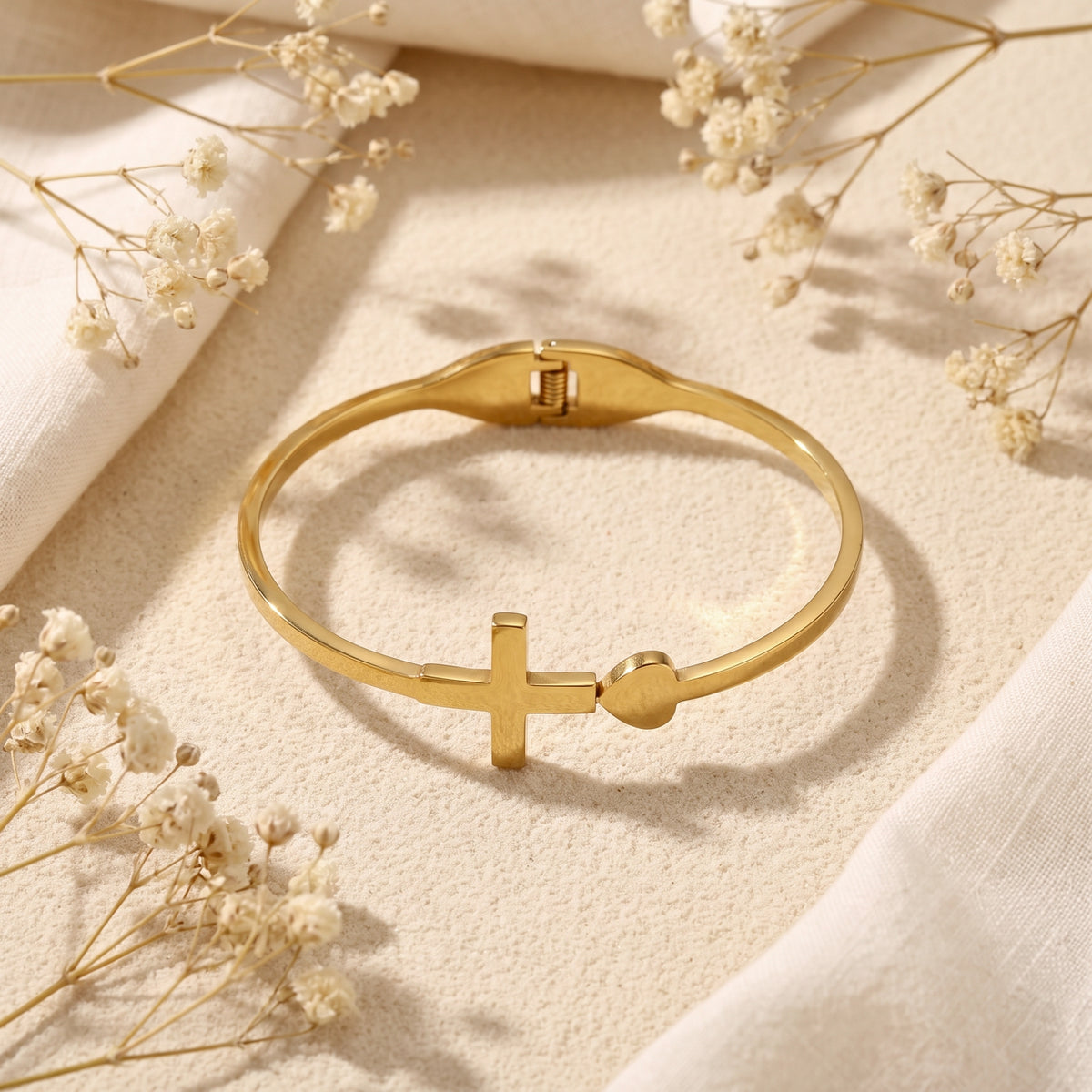 Gold-tone cross and heart hinged bangle bracelet lying flat on beige sand among dried flowers.