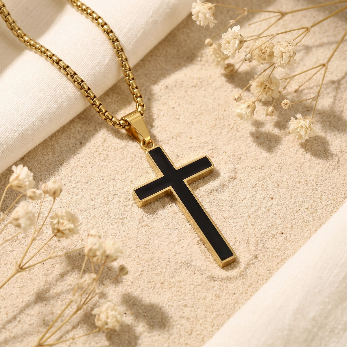 Gold-tone box chain necklace with a black enamel cross pendant, laid on sand with dried flowers.
