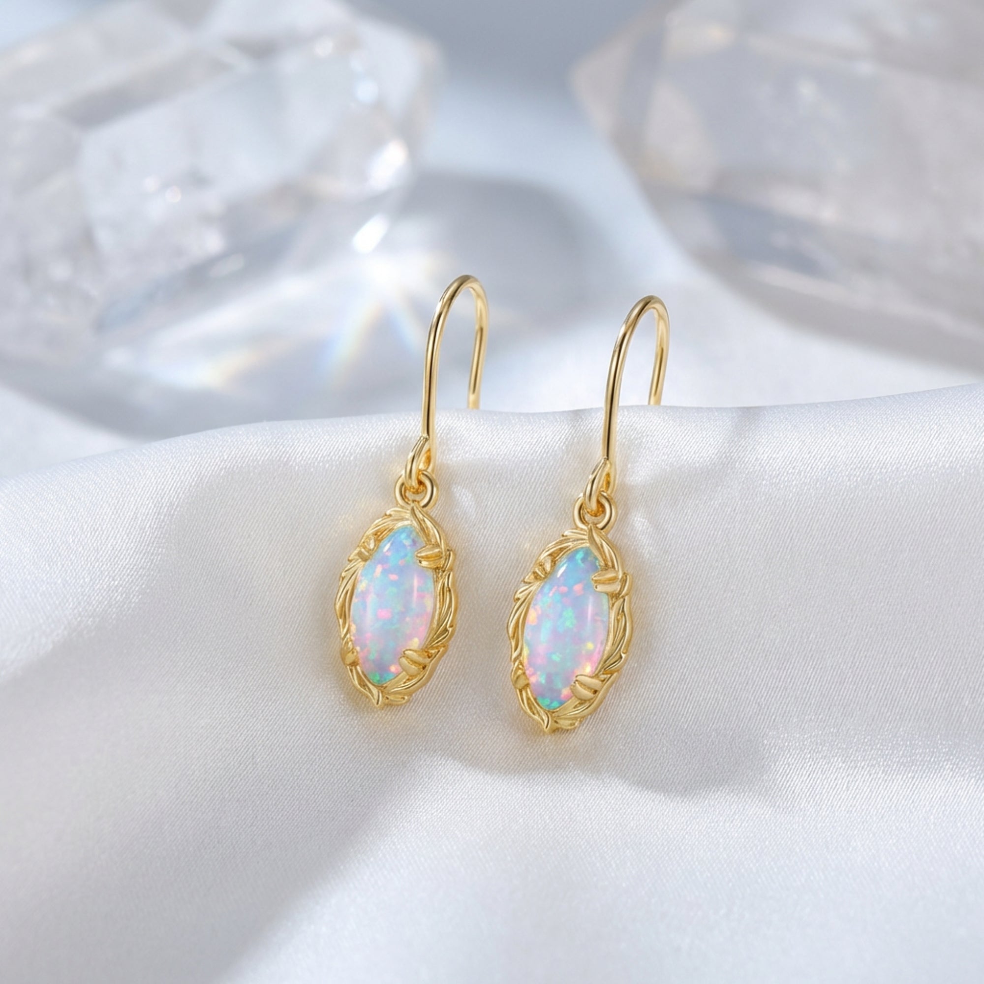 Yellow gold marquise-cut white opal drop earrings styled on white silk fabric with decorative crystals.