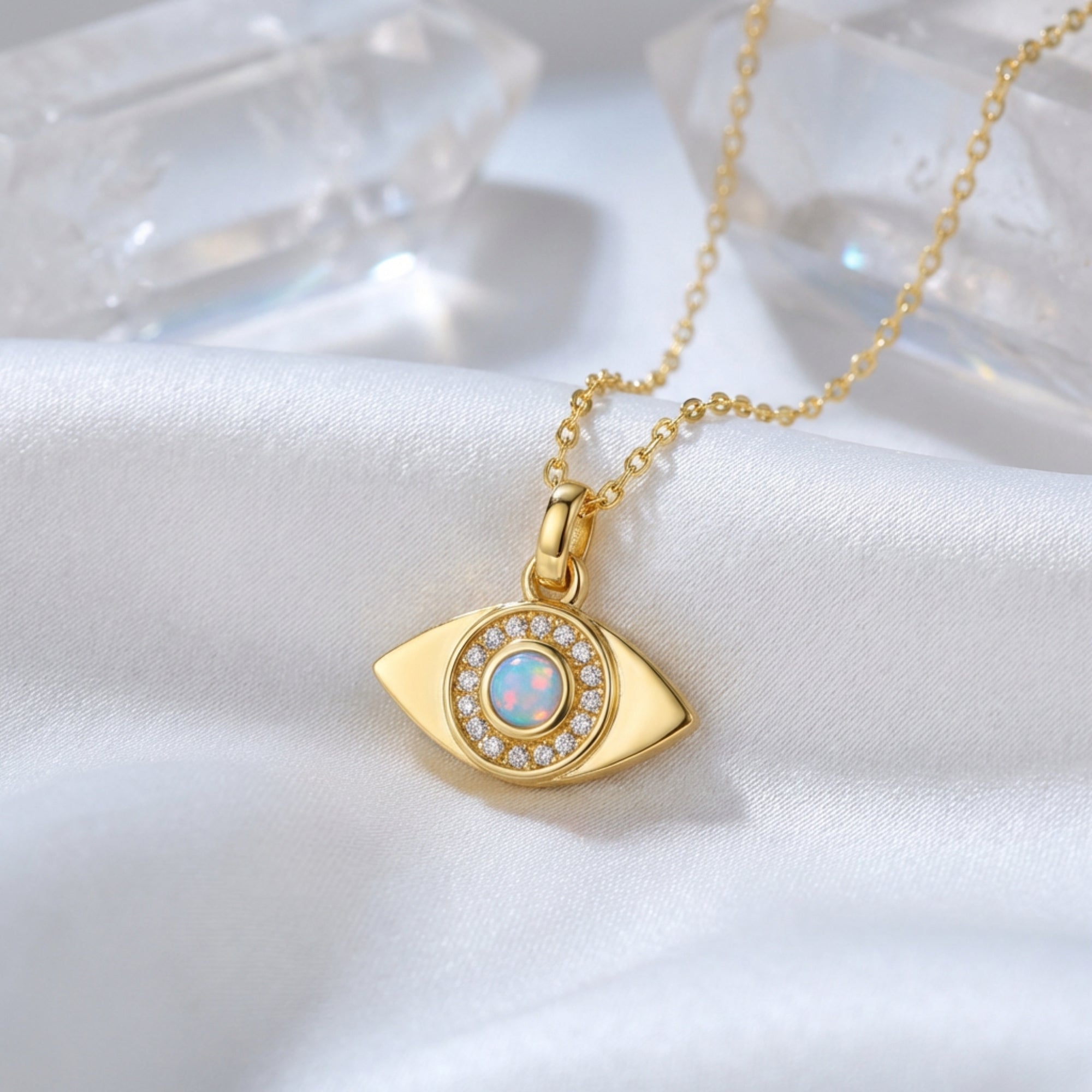 Gold evil eye pendant necklace with a round white opal resting on white silk fabric with crystals.