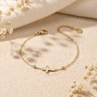 Gold-tone station bracelet with a white cross and bezel-set crystals lying flat on textured sand.