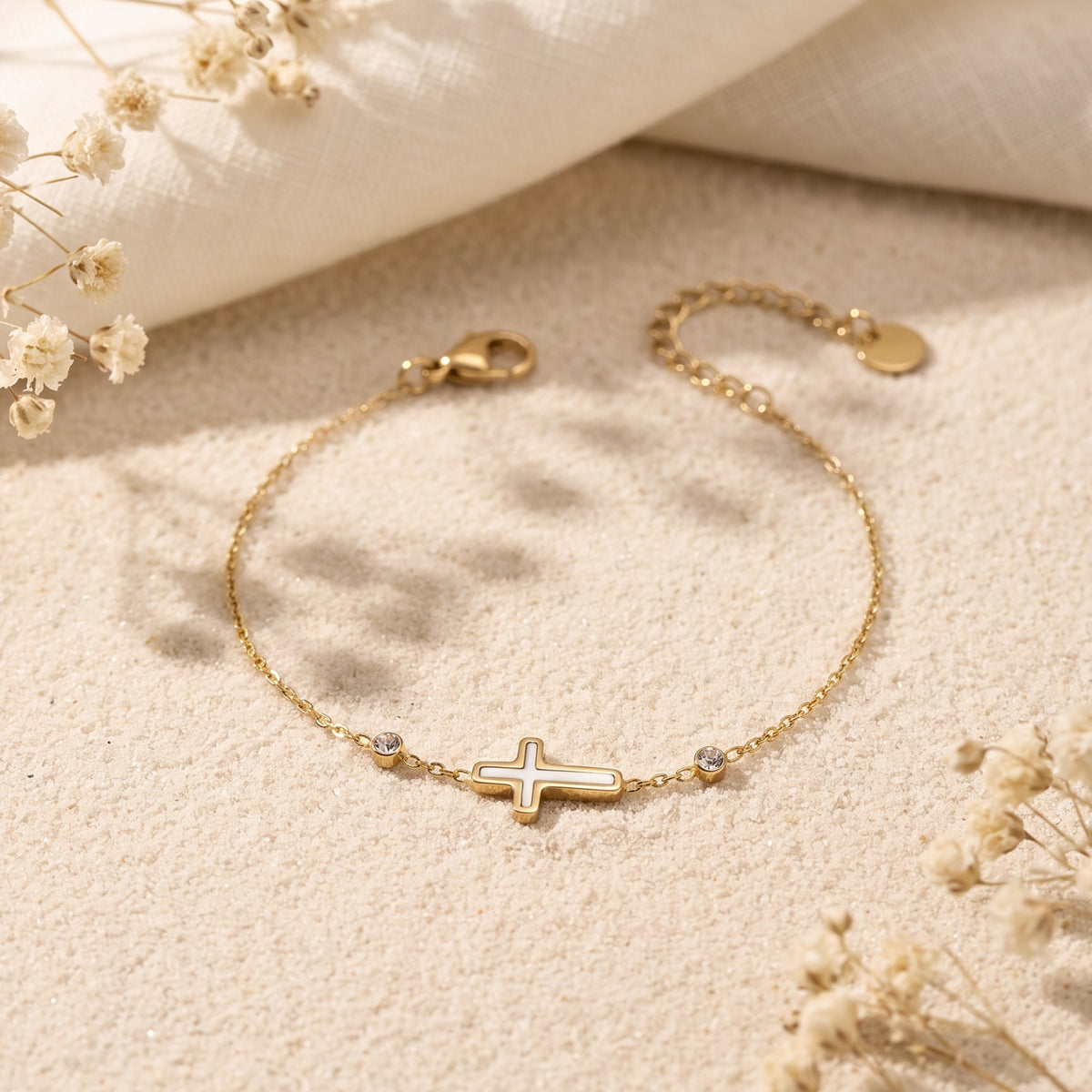Gold-tone station bracelet with a white cross and bezel-set crystals lying flat on textured sand.