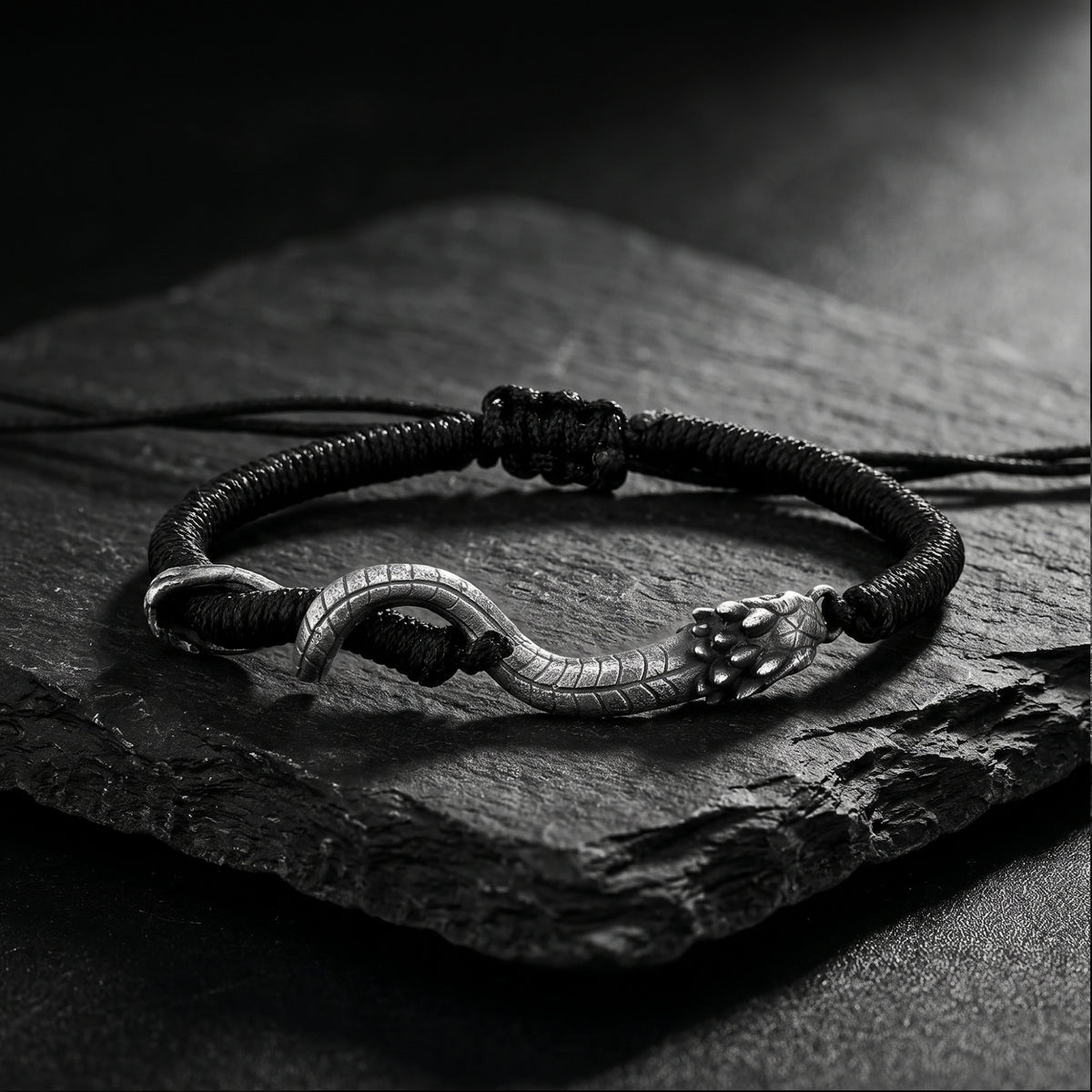 Black cord bracelet with a curved silver serpent centerpiece, arranged on a rough dark stone surface.