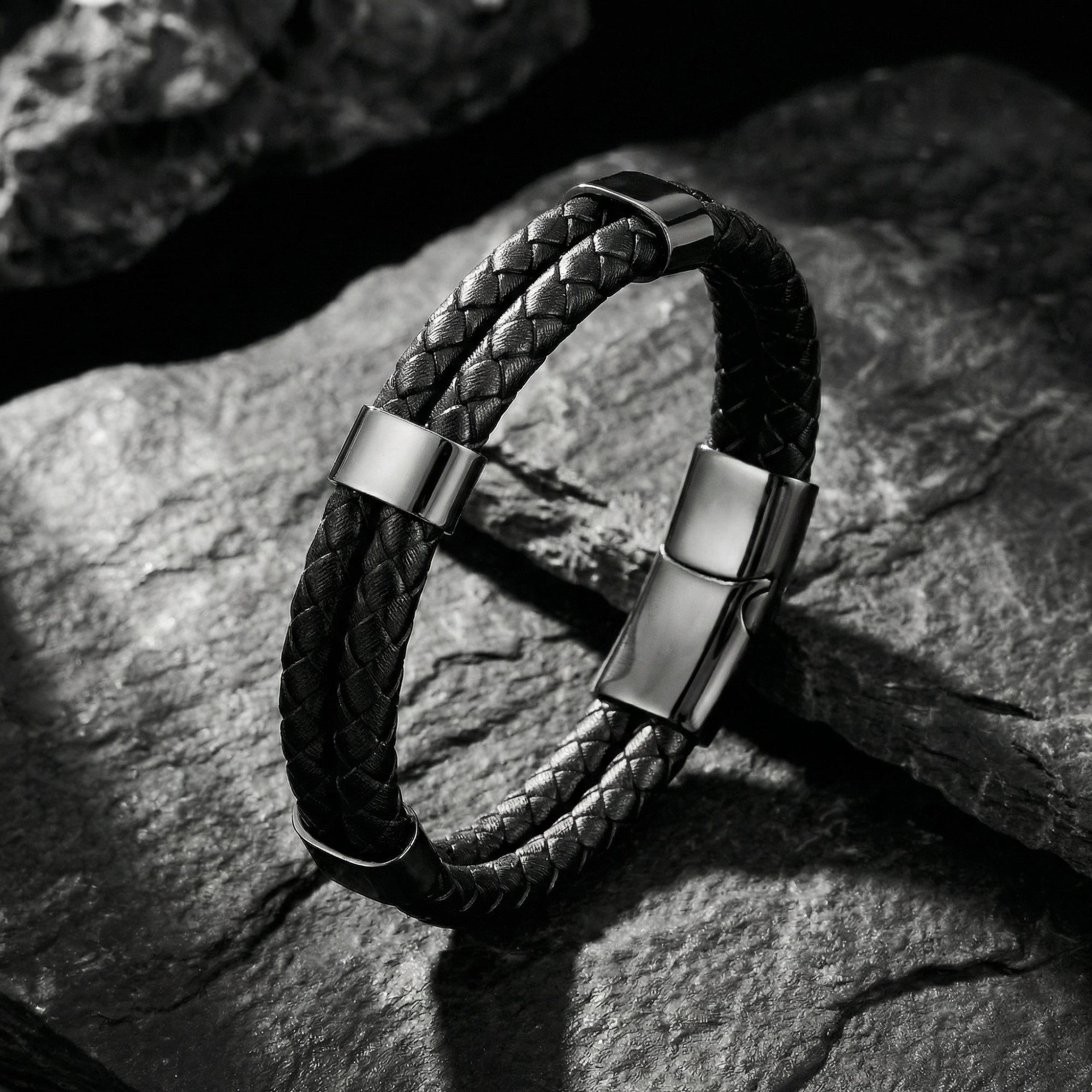 Double-braided black leather bracelet with black stainless steel magnetic clasp resting on dark textured rocks.