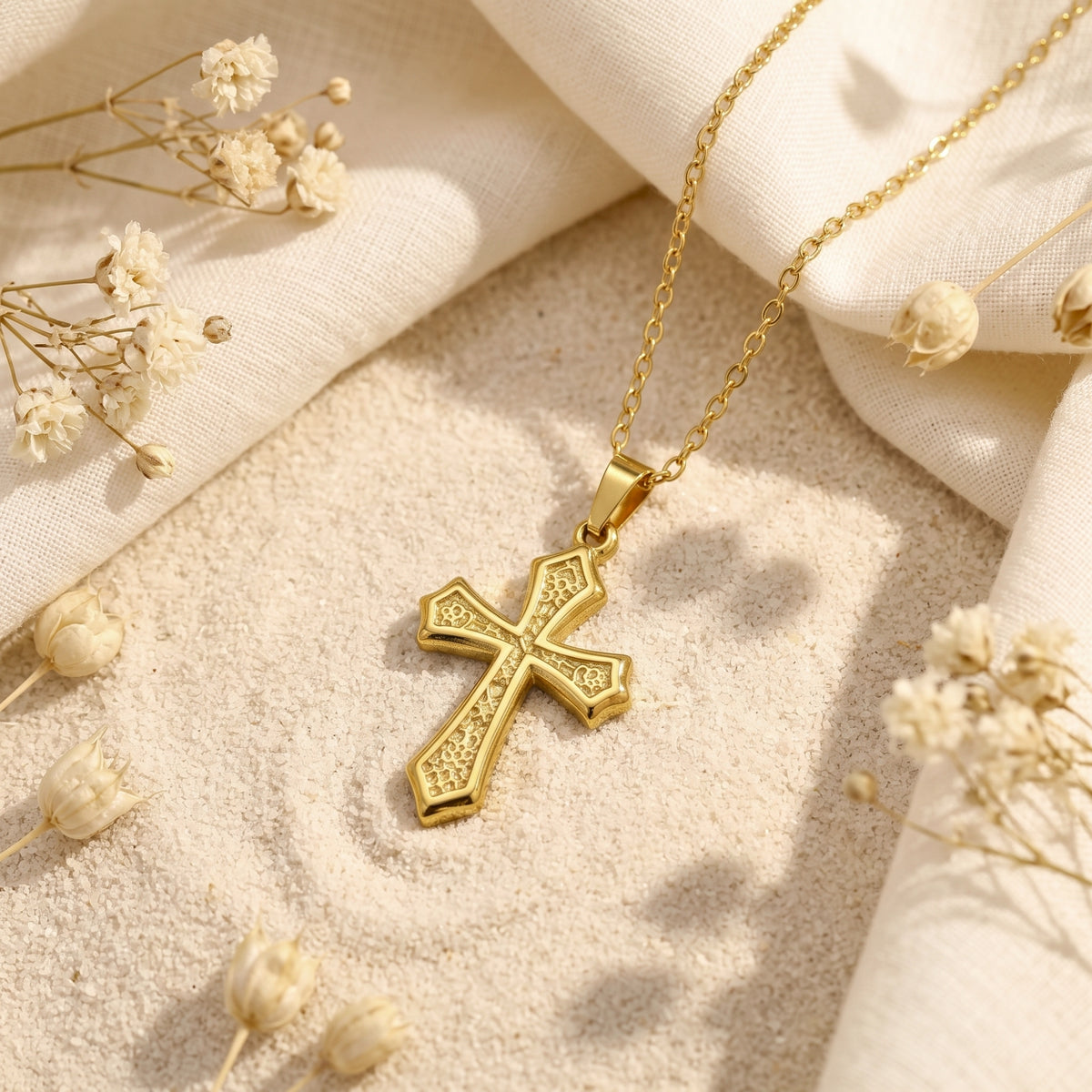 Gold-tone filigree cross necklace resting flat on sand beside white fabric and dried flowers.