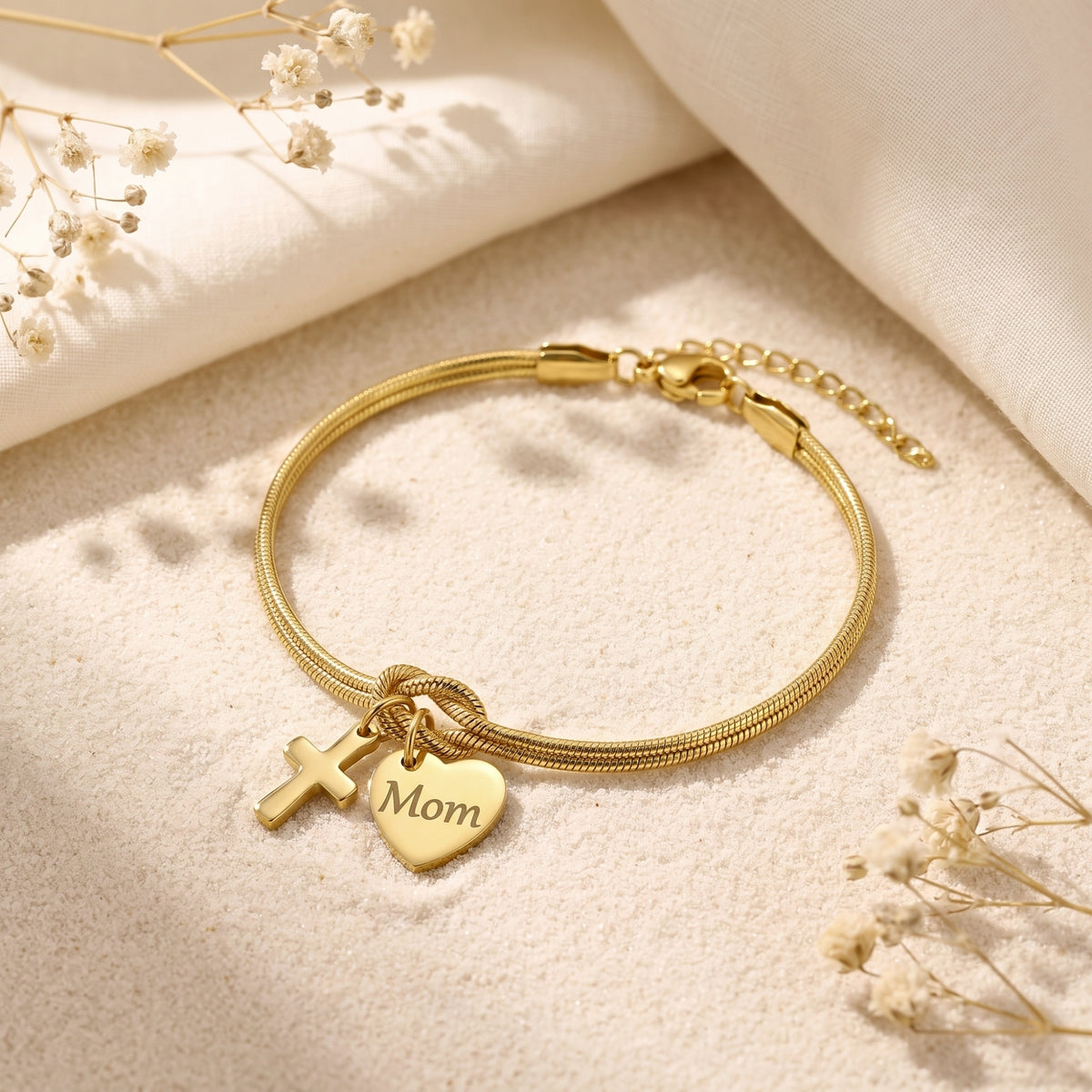 Gold-tone double-strand knot bracelet with a cross and engraved “Mom” heart charm on textured sand.