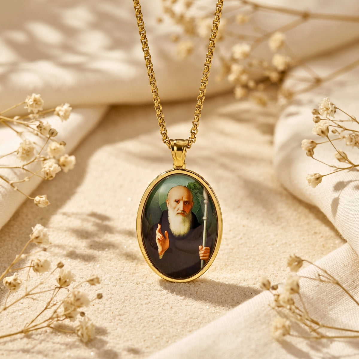 Gold-tone oval saint pendant necklace displayed vertically on sand with white fabric and dried flowers.