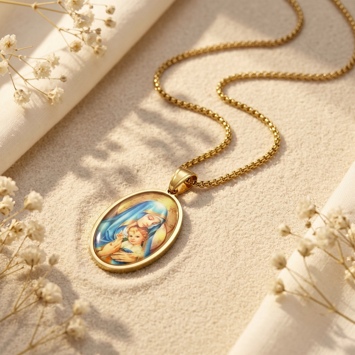 Gold-tone oval Madonna and Child pendant necklace lying on sand with coiled box chain and dried flowers.