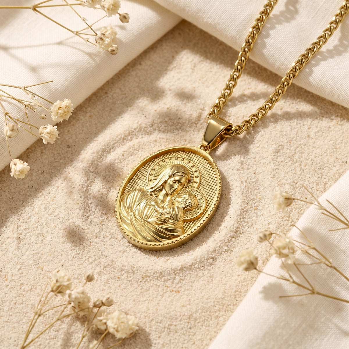 Gold-tone oval Madonna and Child medallion pendant resting on sand with white fabric and dried flowers.