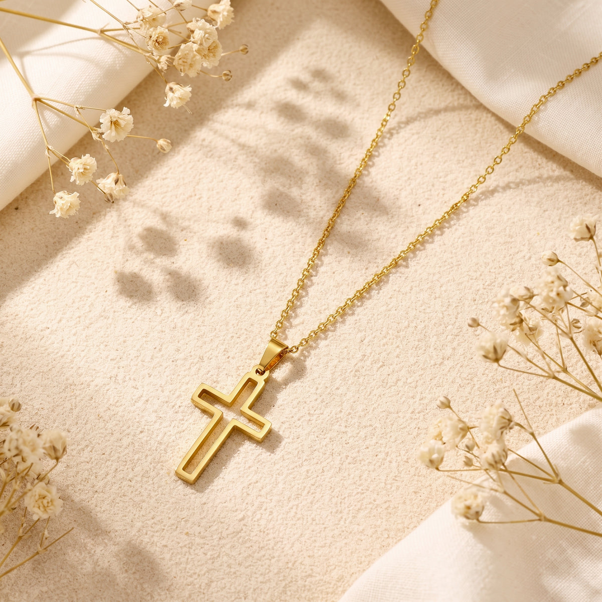 Gold-tone open cutout cross pendant necklace displayed flat on sand with white fabric and dried flowers.