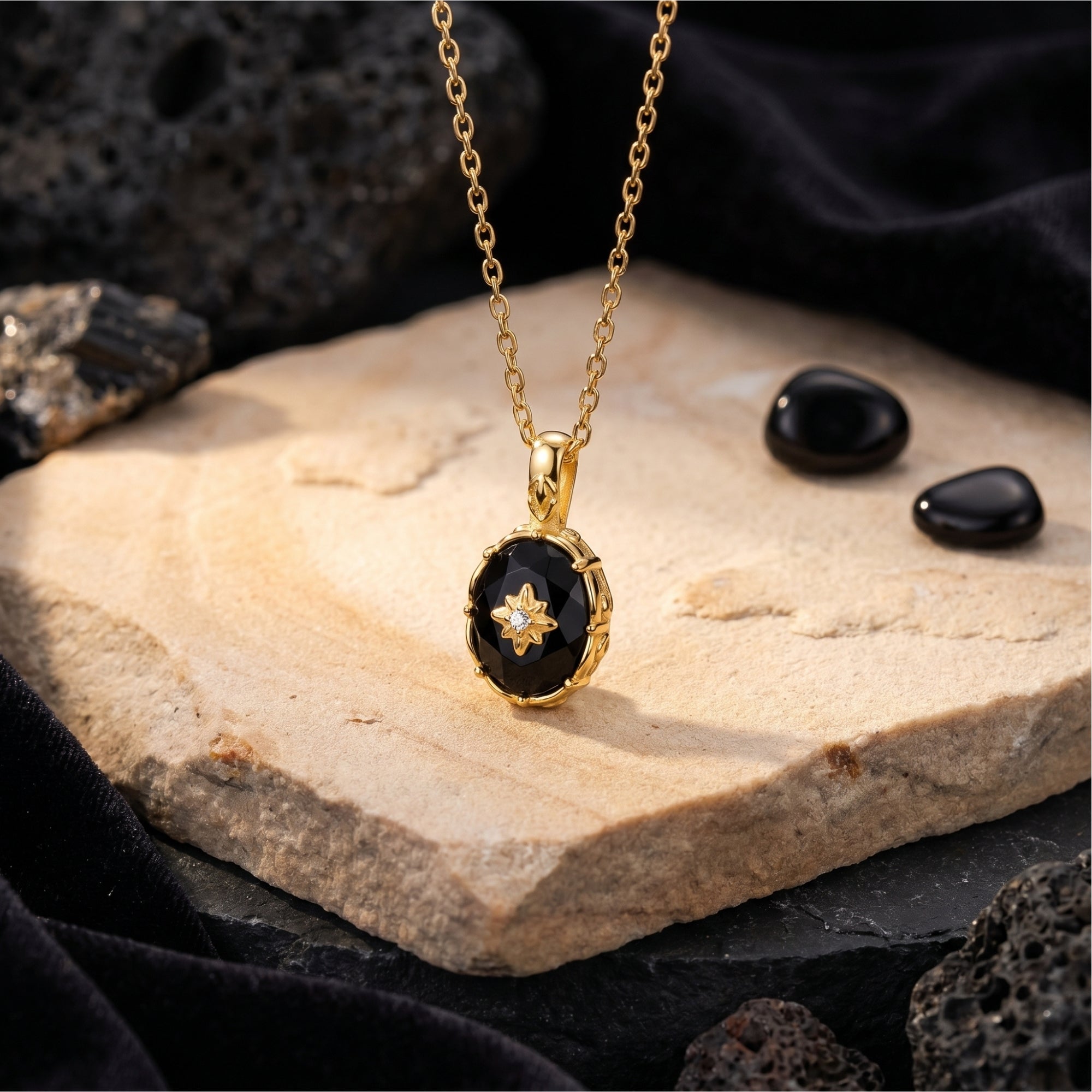 Oval-cut black onyx pendant with a gold starburst on an 18K gold plated chain, styled on a natural stone background.