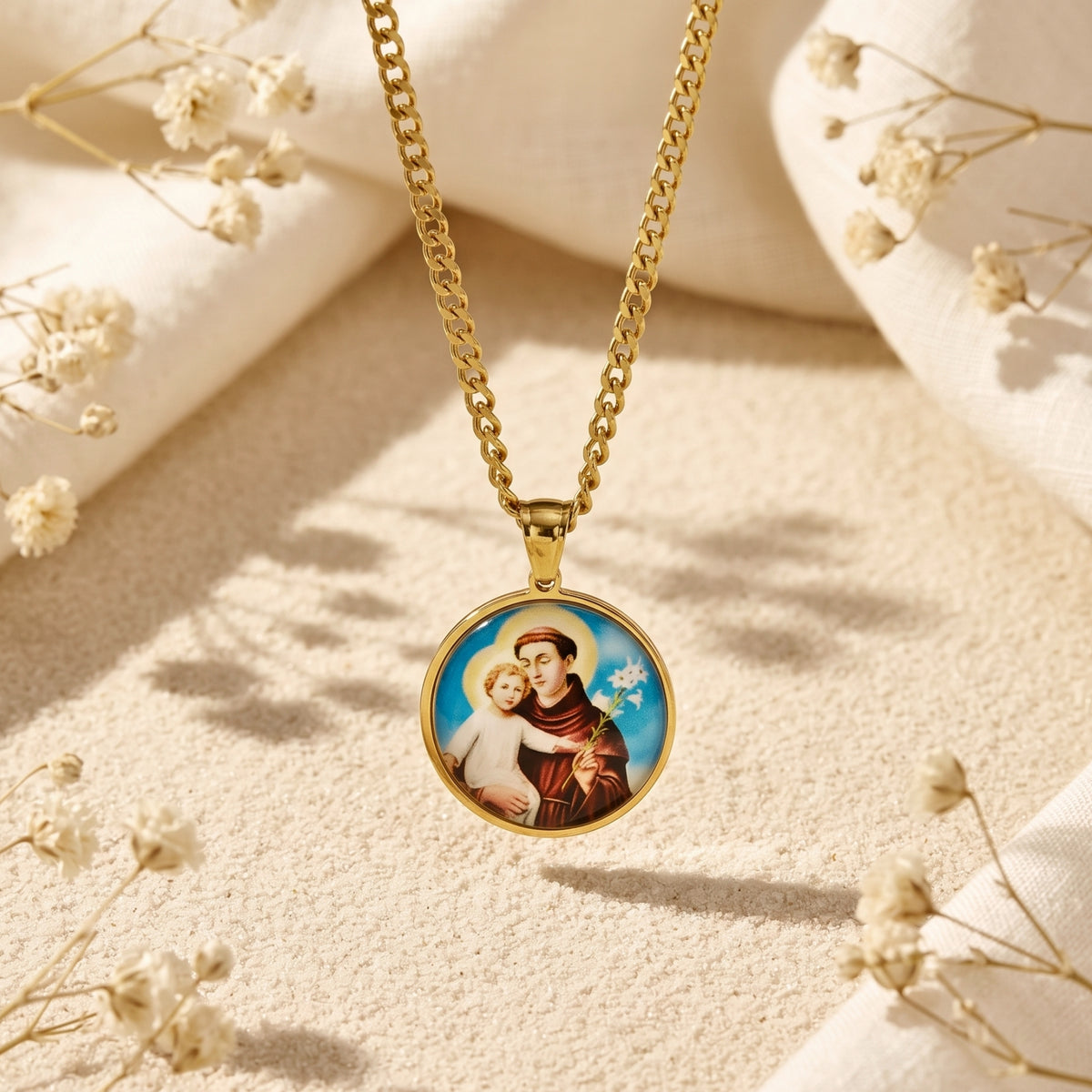 Gold-tone Saint Anthony medallion necklace displayed on sand with white dried flowers and folded neutral fabric.