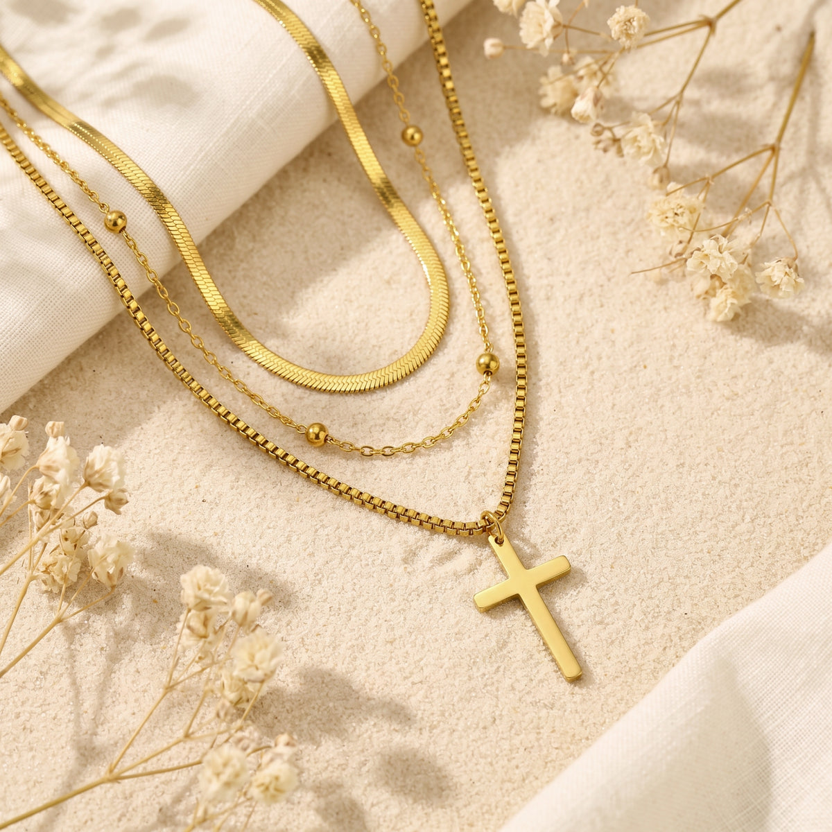 Flat lay of a gold-tone triple-layered cross necklace on sand, linen, and surrounded by white dried flowers.