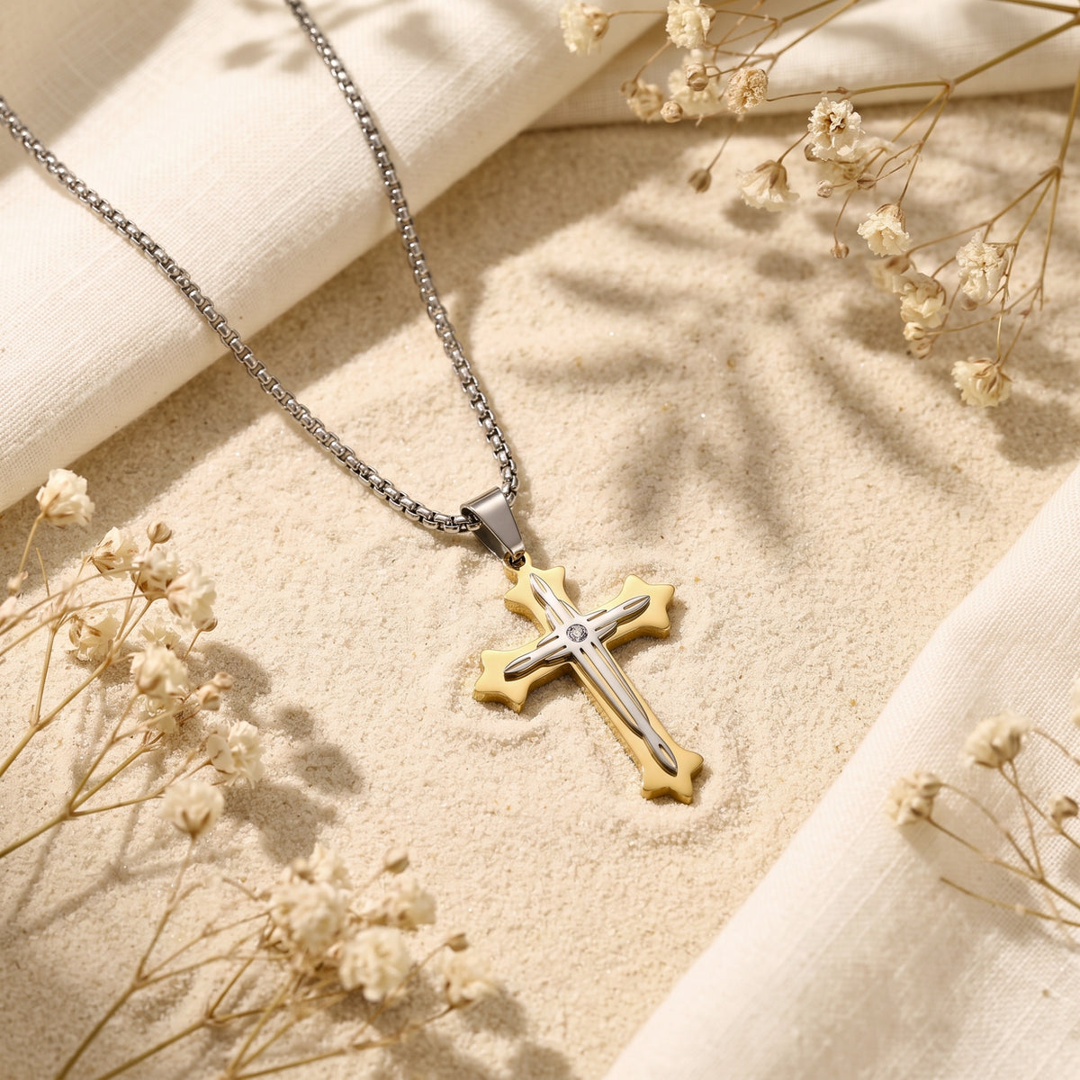 Two-tone cross pendant and chain laid on a sand-colored surface beside white fabric and dried flowers.