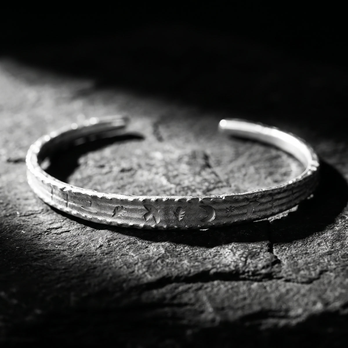Close-up of a silver open cuff bracelet with etched moon, star, and leaf motifs on dark slate.