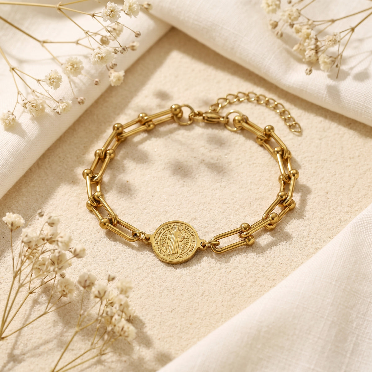 Gold-tone Saint Benedict medallion link bracelet arranged on a textured beige surface with white fabric and dried flowers.