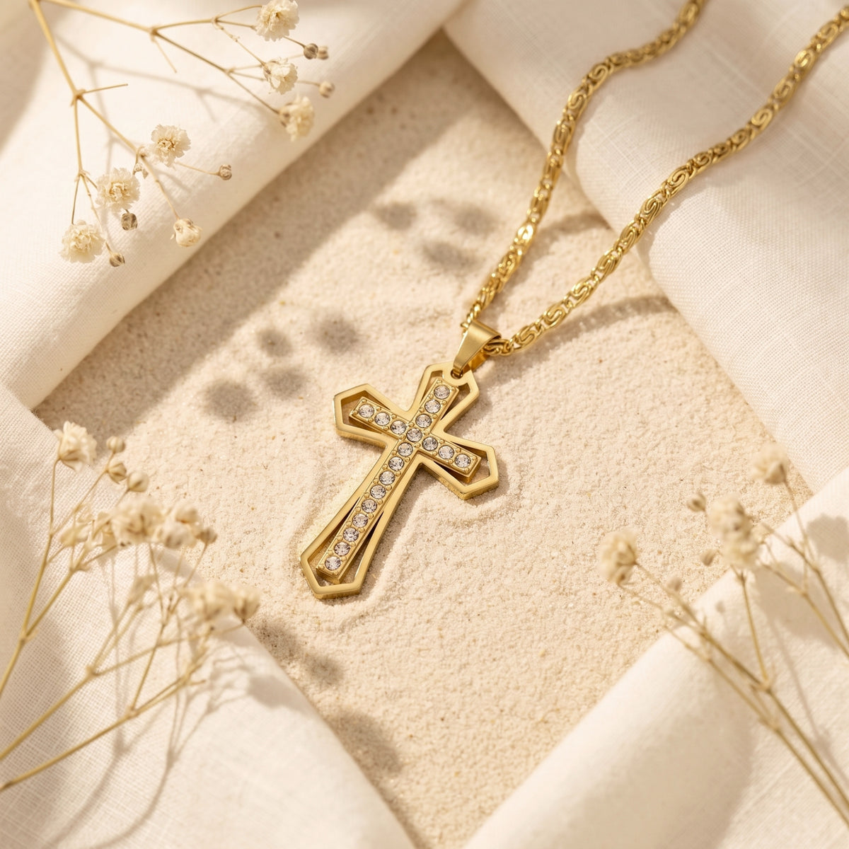 Gold-finish dual-layer cross pendant necklace with clear crystals lying on fine sand with white linen accents.