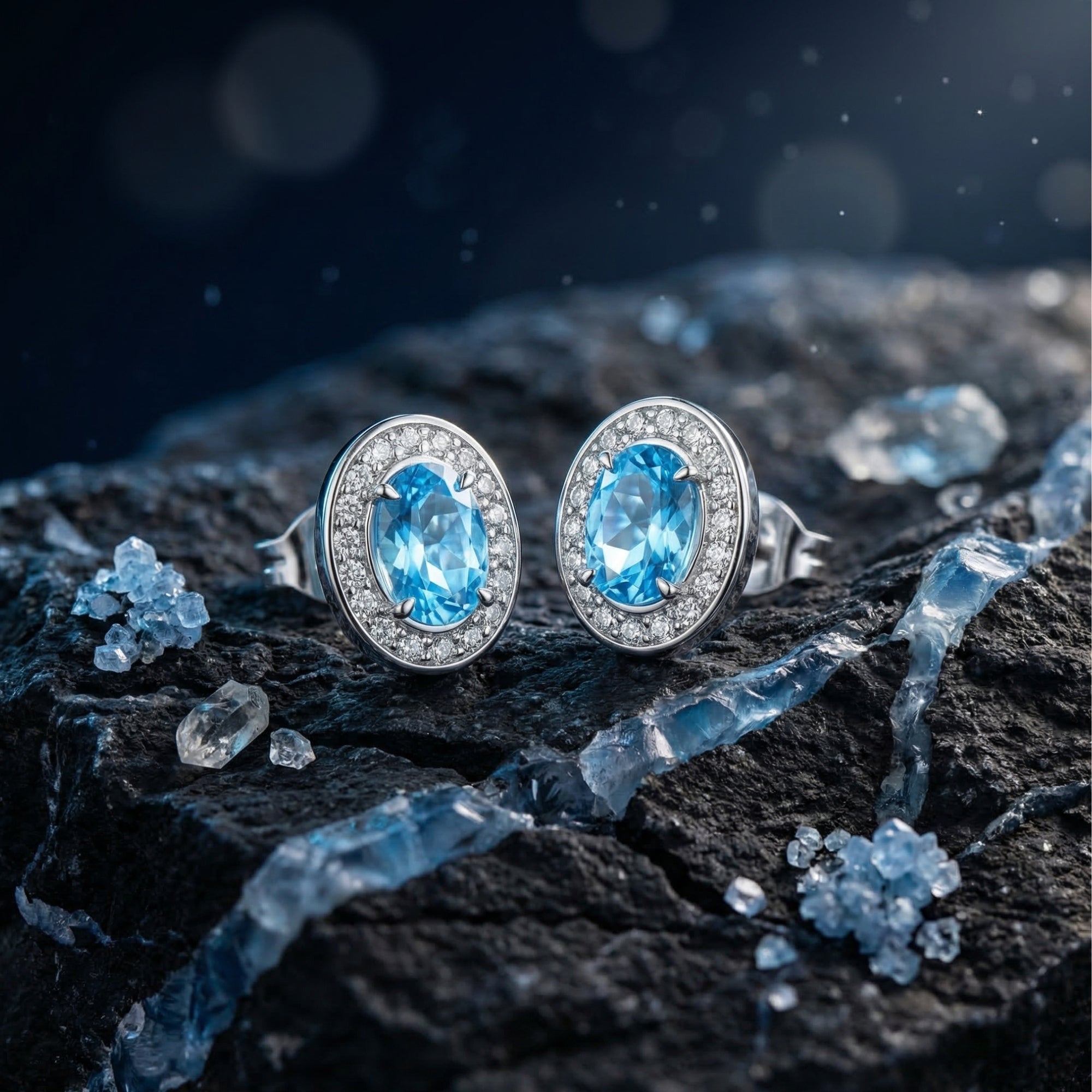 Oval natural blue topaz halo stud earrings in white gold displayed on a dark rock with blue crystals.
