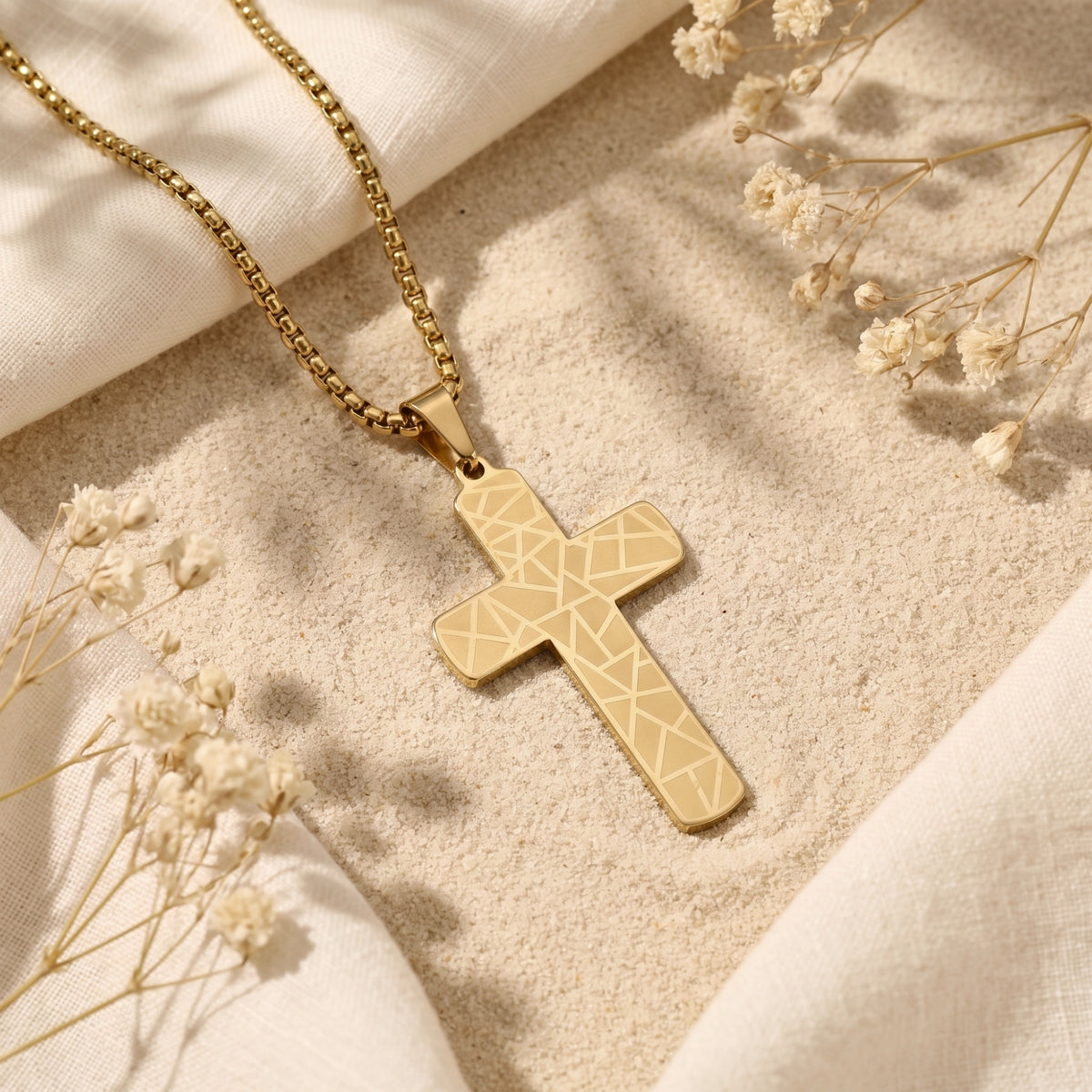 Gold-toned geometric engraved cross pendant on a box chain, laid flat on sand with white cloth and dried flowers.