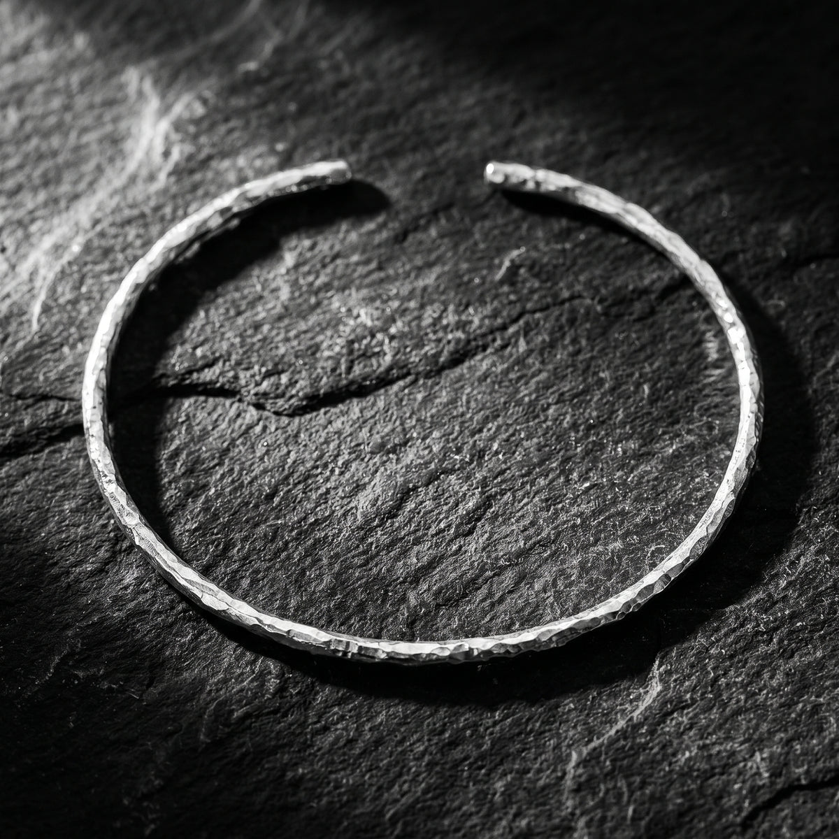 Top-down view of a slim hammered silver open cuff bracelet resting on dark stone.