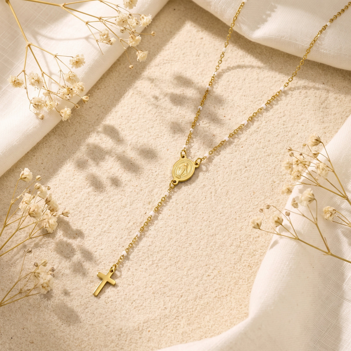 Gold-tone rosary lariat necklace lying on sand with dried white flowers, an oval medal, and a cross drop.