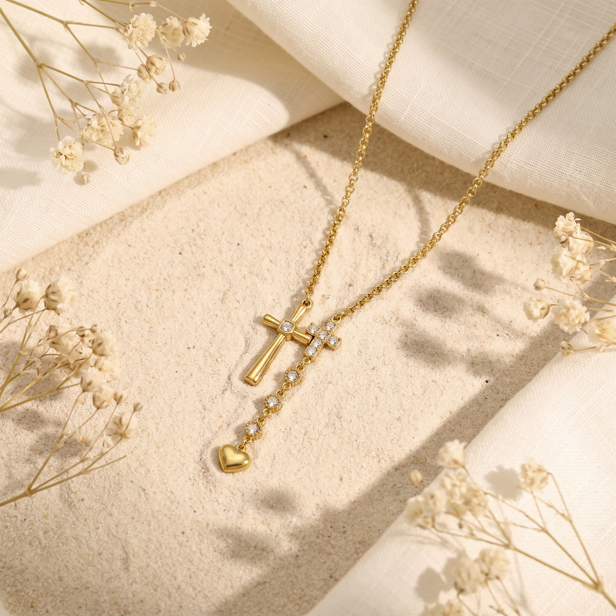 Gold double cross lariat necklace lying on sand with draped white cloth and dried flowers.