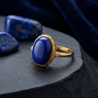Oval lapis lazuli ring in yellow gold , styled on a dark stone surface with raw lapis lazuli gemstones.