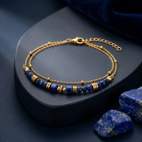 18K gold plated double chain bracelet with natural lapis lazuli beads on a dark pedestal with raw stones.