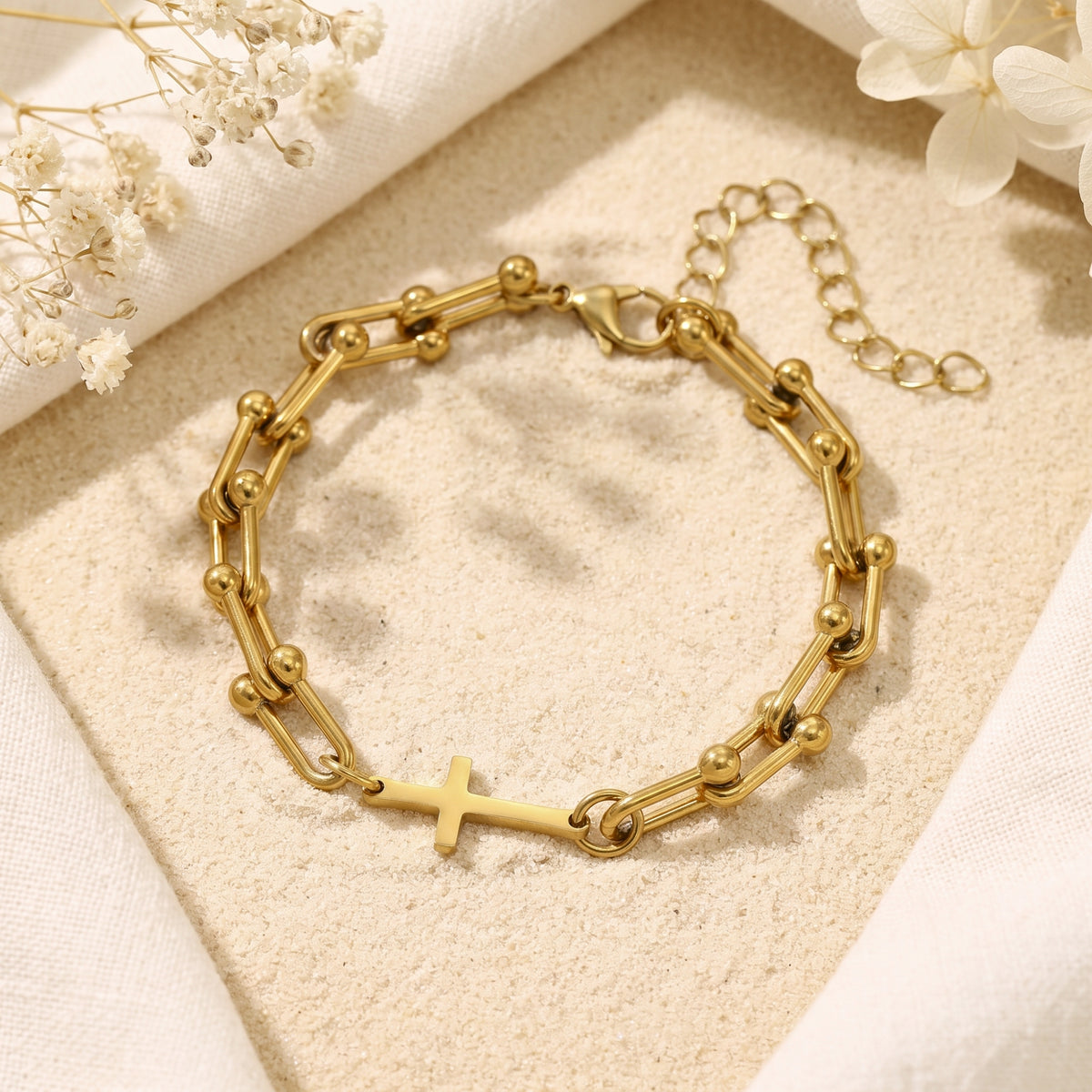 Gold-tone U-link bracelet with an integrated cross, displayed flat on sand with white fabric and dried flowers.