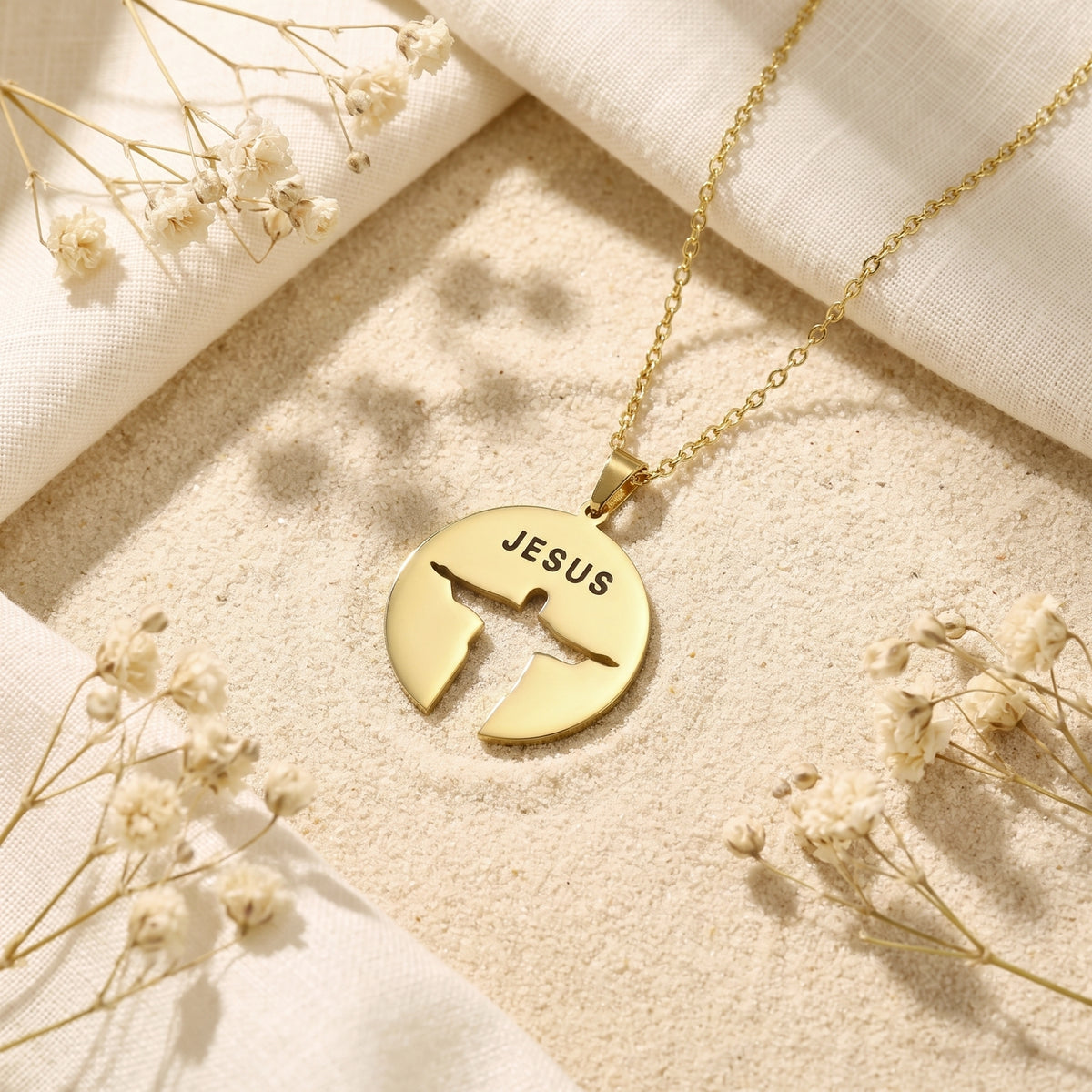Gold-tone disc necklace with JESUS text and a Christ silhouette cutout on sand among dried flowers.