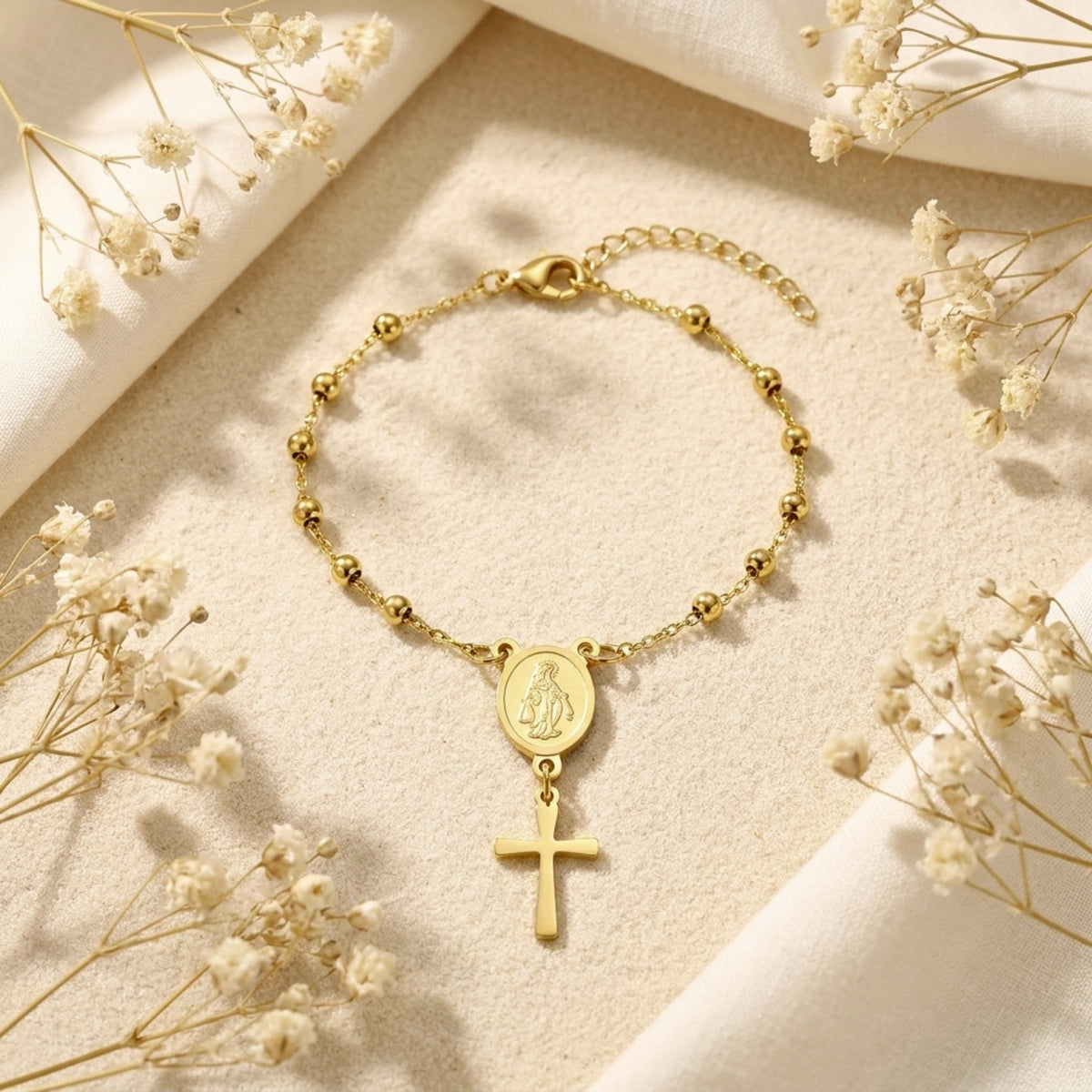 Gold-tone rosary bracelet with round beads, Virgin Mary medal, and cross charm on beige fabric with dried flowers.