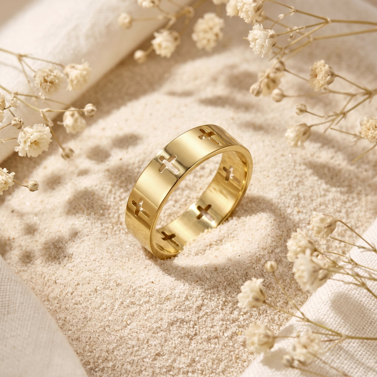 Gold-tone band ring with cutout cross motifs angled on fine sand near white dried flowers.